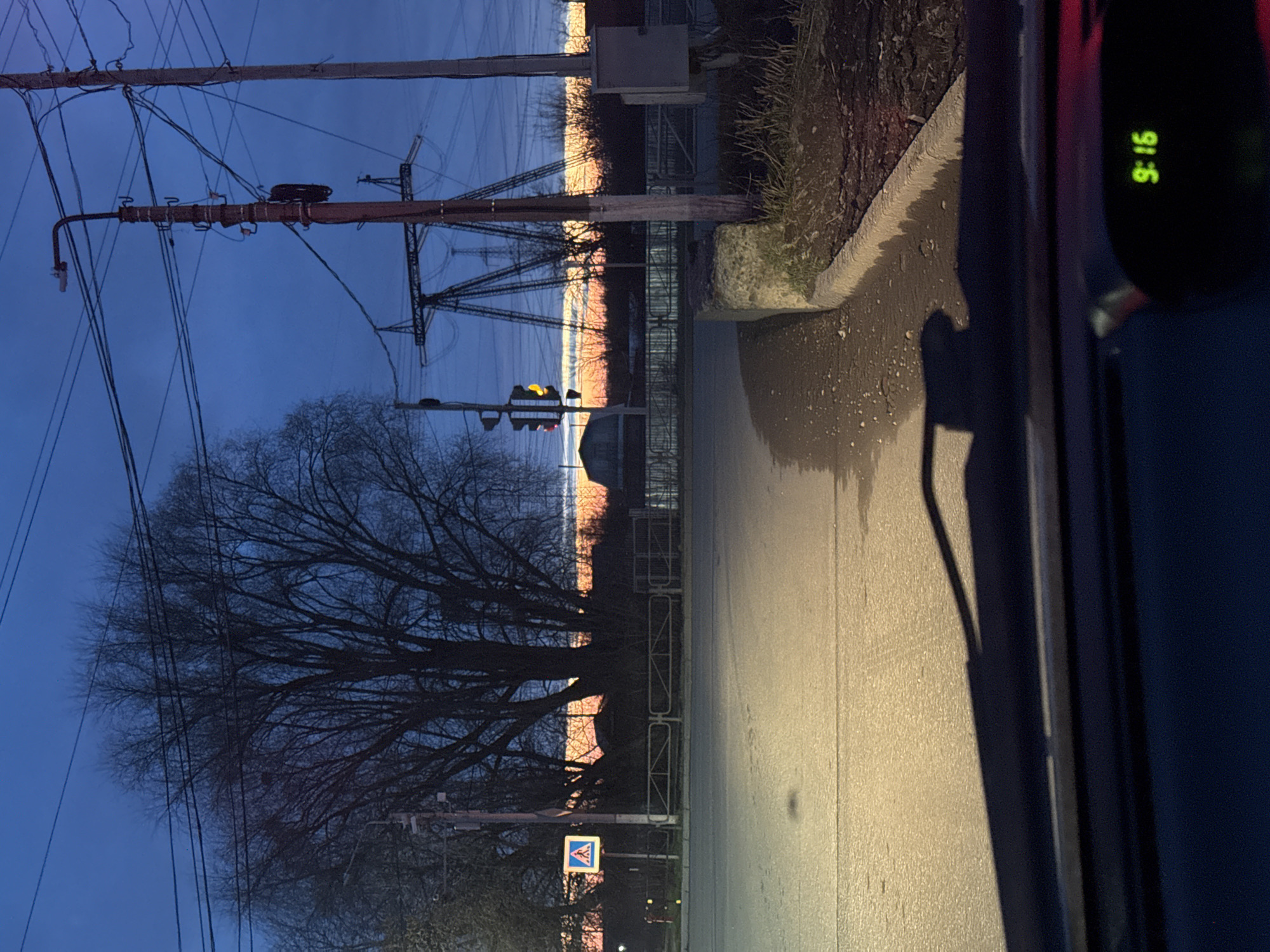 The image depicts a street scene at twilight, possibly in Chaikovsky, Russia.
**Environment:**
* A paved road is visible, appearing wet, possibly due to recent rain.
* A curb separates the road from a grassy area.
* Overhead power lines and electrical pylons dominate the background.
* Bare trees stand alongside the road.
**Objects:**
* A traffic light is visible in the middle distance.
* A crosswalk sign is present near the bottom of the image.
* A car's interior is partially visible on the right side of the image, including a digital clock displaying "9:16".
**Time:**
* The time is 9:16 pm, as displayed on the car's clock.
* The twilight sky and street lighting suggest it is evening.
**People/Actions:**
* There are no people visible in the immediate scene.
* The image is taken from inside a car.
**Possible Location:**
* Given the description of the city's features, the presence of overhead electrical infrastructure, trees, and the surrounding environment, this may be a road near an industrial area in Chaikovsky, Russia.