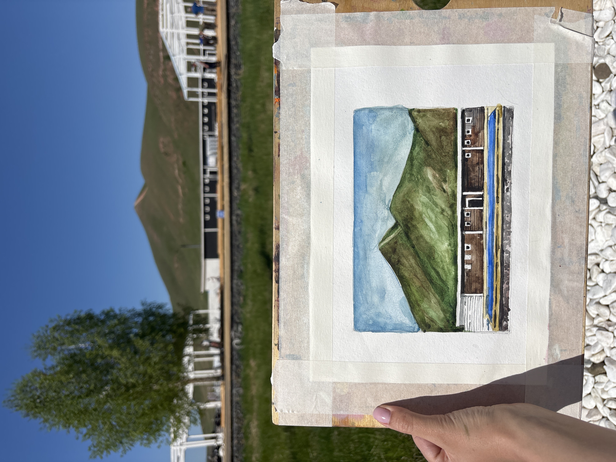 A hand holds up a small watercolor painting, taped to a wooden board, against a vibrant outdoor backdrop in Дрокино, Russia. The painting itself features a stylized landscape: a large green mountain occupies the center-left, with a light blue expanse above and to its left, possibly depicting the sky or a body of water. To the right of the mountain are rectangular architectural elements, including structures with brown facades and small white windows, a prominent vertical blue section, and details in grey and yellow.
The background behind the painting reveals a clear, bright blue sky in the upper left. Gently undulating green hills or mountains stretch across the horizon. Below these, and predominantly in the mid-ground, are modern-looking white structures, likely pergolas or sections of a building, with blurred figures of people discernible within them. A lush green tree stands blurred in the lower left background. To the right of the painting, the ground is covered in white gravel or stones.
The scene captures a moment of creative activity, likely plein air painting, or the presentation of a newly finished artwork within the environment that inspired it. The strong sunlight and clear sky indicate a bright day. No discernible text is visible within the image.