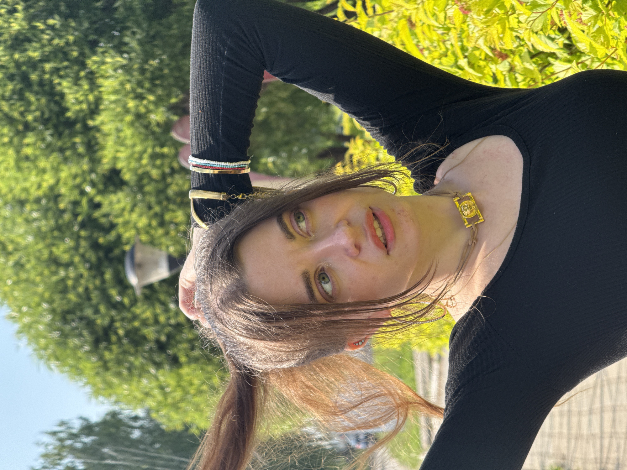 A medium shot captures a young woman outdoors, her body angled to the right and her head turned to her left, looking away from the camera. She has long, brown hair that is being pulled up into a ponytail. Her left hand is in her hair, while her right arm is bent at the elbow and raised behind her head, resting on her arm. She wears a black, long-sleeved, ribbed shirt. Around her neck is a choker necklace with a rectangular gold pendant featuring a circular emblem. On her right wrist are several beaded bracelets. The background is a blur of green foliage and a bright blue sky, suggesting an outdoor, park-like setting. The lighting indicates it is daytime with clear weather.