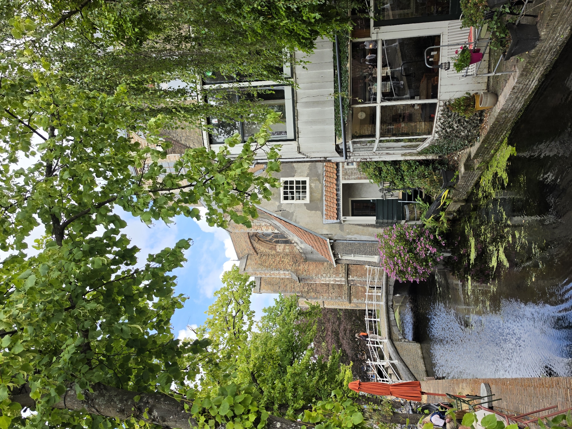 This image depicts a picturesque canal scene in Delft, Netherlands. The canal, reflecting the overcast sky and surrounding greenery, flows through a narrow urban waterway. A white arched bridge crosses the canal, with a person standing on it, facing away from the camera and looking towards a large, old brick building with arched windows that appears to be a church or historical structure. On the right side of the canal, a multi-story building with large windows, some of which are reflective, is visible. The lower part of this building has a glass enclosure, suggesting it might be a cafe or restaurant with outdoor seating. Potted plants and greenery are scattered along the canal banks and around the buildings. An orange umbrella is unfurled near the left bank, adjacent to seating and a table. The overall atmosphere is serene and historic, characteristic of Dutch canal towns.
