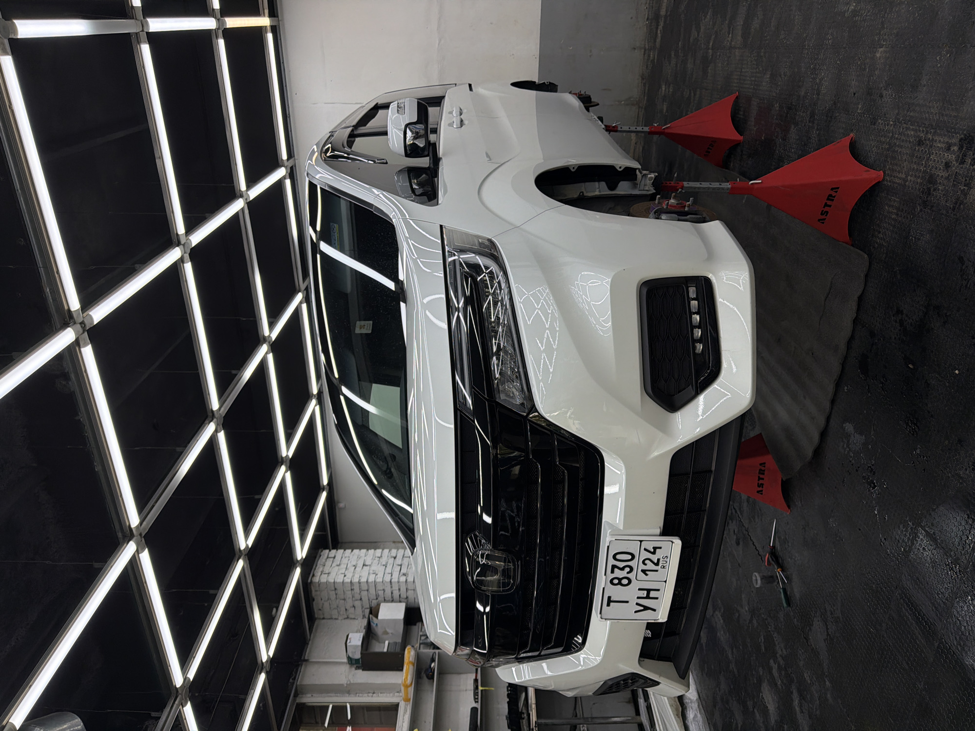 A white Honda Stepwgn minivan is parked in an indoor garage, possibly a car repair shop or detailing studio, in Krasnoyarsk, Russia. The vehicle is raised on four red jack stands, indicating it is undergoing maintenance or inspection. The garage has a dark ceiling with an array of bright, grid-like fluorescent lights. The floor is black and textured. Tools, including screwdrivers, are visible on the floor near the front of the vehicle. The license plate on the front of the car reads "T 830 YH 124 RUS". The minivan's front grille, headlights, and bumper are clearly visible, as are the side mirrors and doors. The overall impression is of a professional automotive service environment.