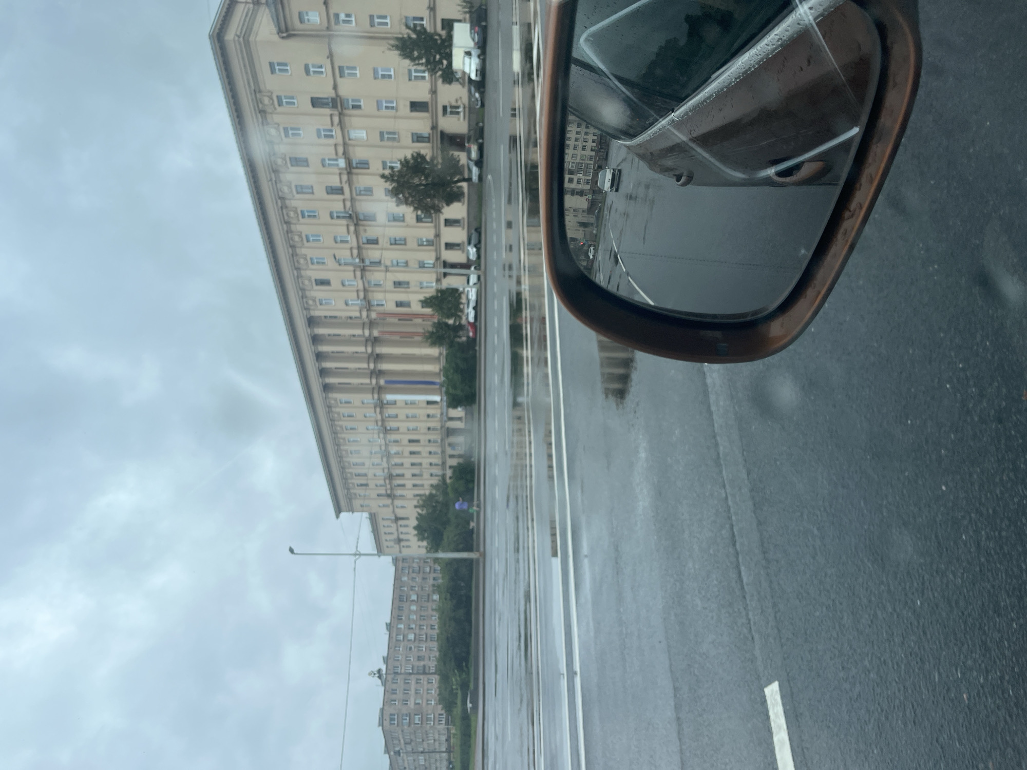 The image captures a cloudy day in Saint Petersburg, Russia, likely on a street with a wide road. In the foreground, a car's side-view mirror is prominently featured, reflecting the street scene behind the vehicle. The road is wet, with puddles indicating recent rain. Beyond the road, a large, beige, multi-story building dominates the background. This building features a classical architectural style with numerous windows and columns. To the left of this main building, another, less ornate, multi-story residential building is visible. Several cars are parked along the street in front of these buildings. The sky is overcast with a pale, diffused light. In the reflection of the side-view mirror, a silver car is seen driving on the wet road, heading away from the viewer. The overall scene conveys a sense of urban life on a damp, overcast day.