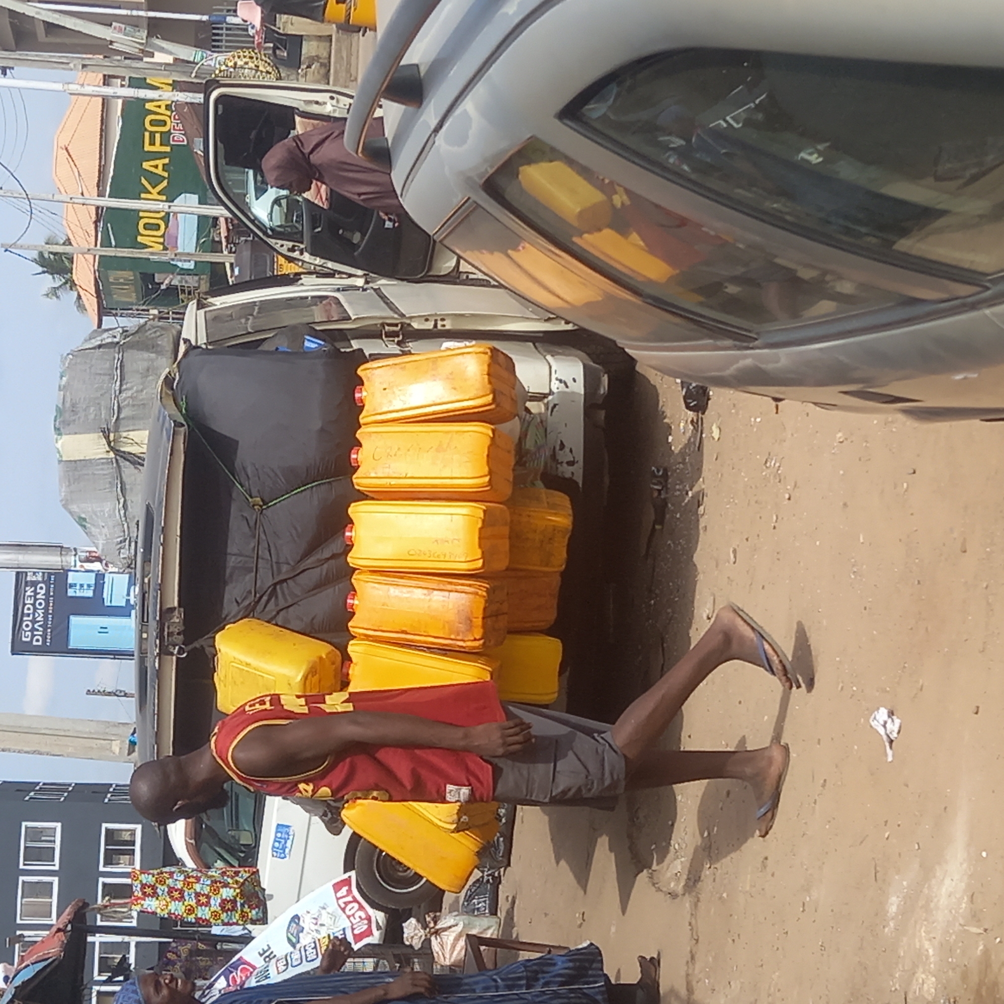The image shows a street scene in what appears to be Dadi Riba, Nigeria, based on the text on the side of a building visible in the background. A man wearing a red tank top and gray shorts is standing on the side of a white truck. The truck is loaded with stacked yellow containers. Another vehicle, light gray in color, is parked to the right of the truck, partially obstructing the view. In the background, there is a building with a green sign that reads "MOUKA FOAM" in large letters. A billboard with the words "GOLDEN DIAMOND" is visible in the distance. There are also people standing on the side of the street. The ground is dusty and sandy.