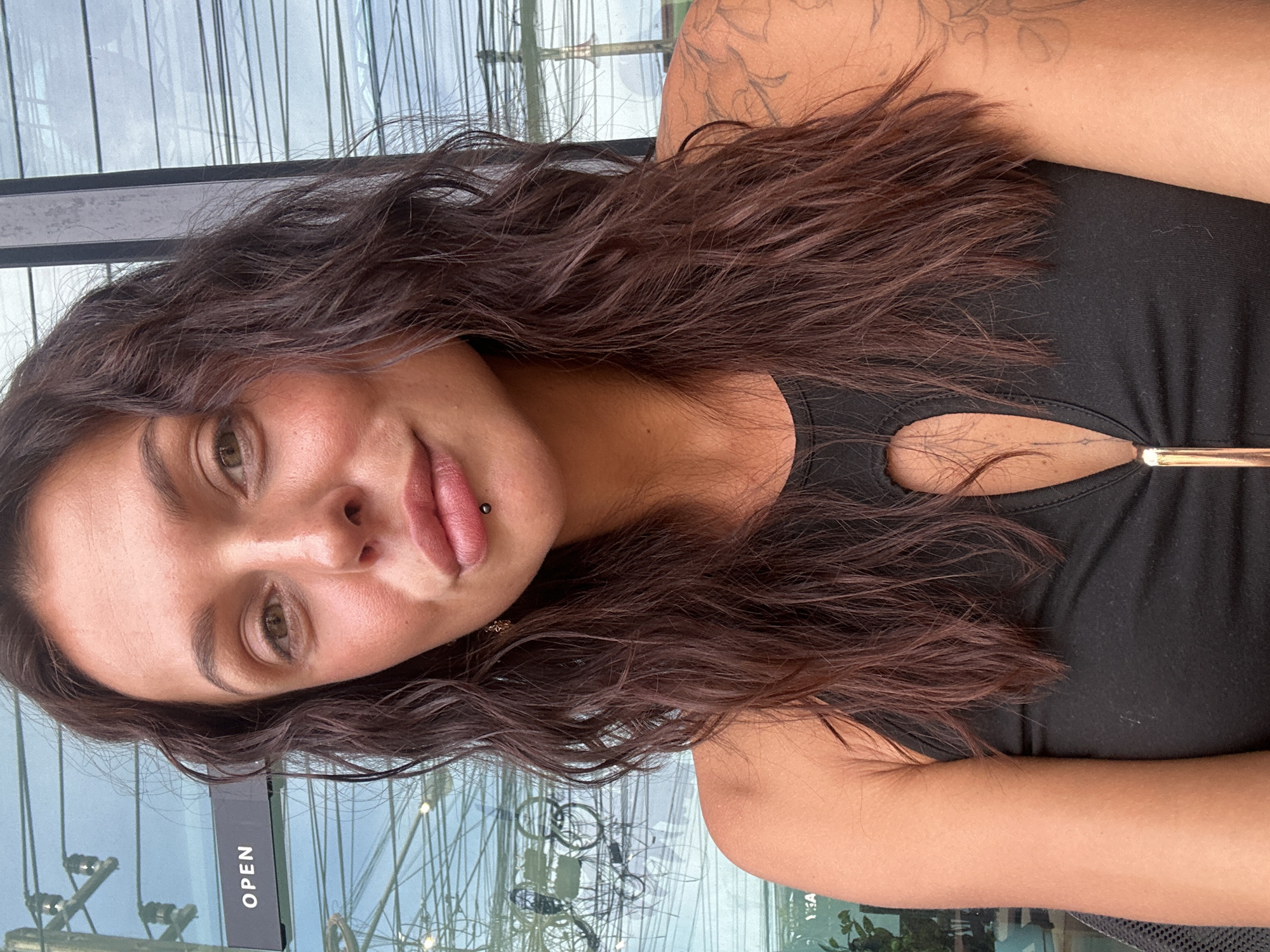 A medium shot selfie captures a young woman with long, dark, wavy hair and fair skin. She has light brown eyes, full lips with a dark stud piercing in the center, and a hint of blush on her cheeks. She is wearing a black tank top with a keyhole cutout at the chest. A portion of a tattoo is visible on her right shoulder and upper arm. Behind her, a glass window reflects a street scene with a sign that reads "OPEN." The location is identified as a street in the city of Ko Samui District, Thailand.