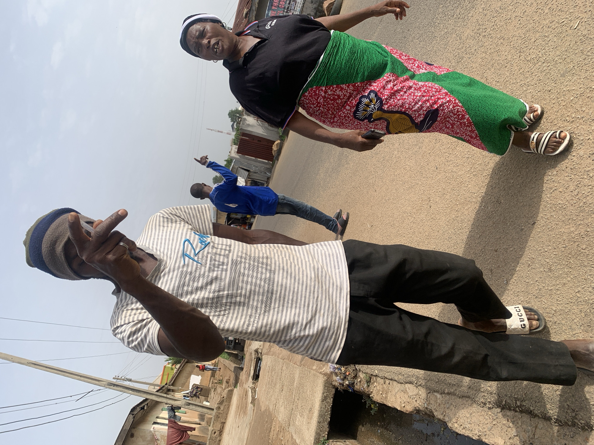 The image shows three people on a street in what appears to be a residential or commercial area.
A person in the foreground, possibly male, wears a striped shirt, black pants, and slides. They are holding their hand up near their face.
Behind them, a person in a blue shirt and jeans gestures with their right hand.
Further behind and to the right, a person wearing a black shirt and a patterned skirt, possibly female, stands in the street. They are holding a dark-colored object, possibly a phone, in their hand.
The street appears to be paved, with a concrete curb and drainage ditch along the side. Buildings and power lines are visible in the background.
The location is suspected to be in the city of Jos, Nigeria.