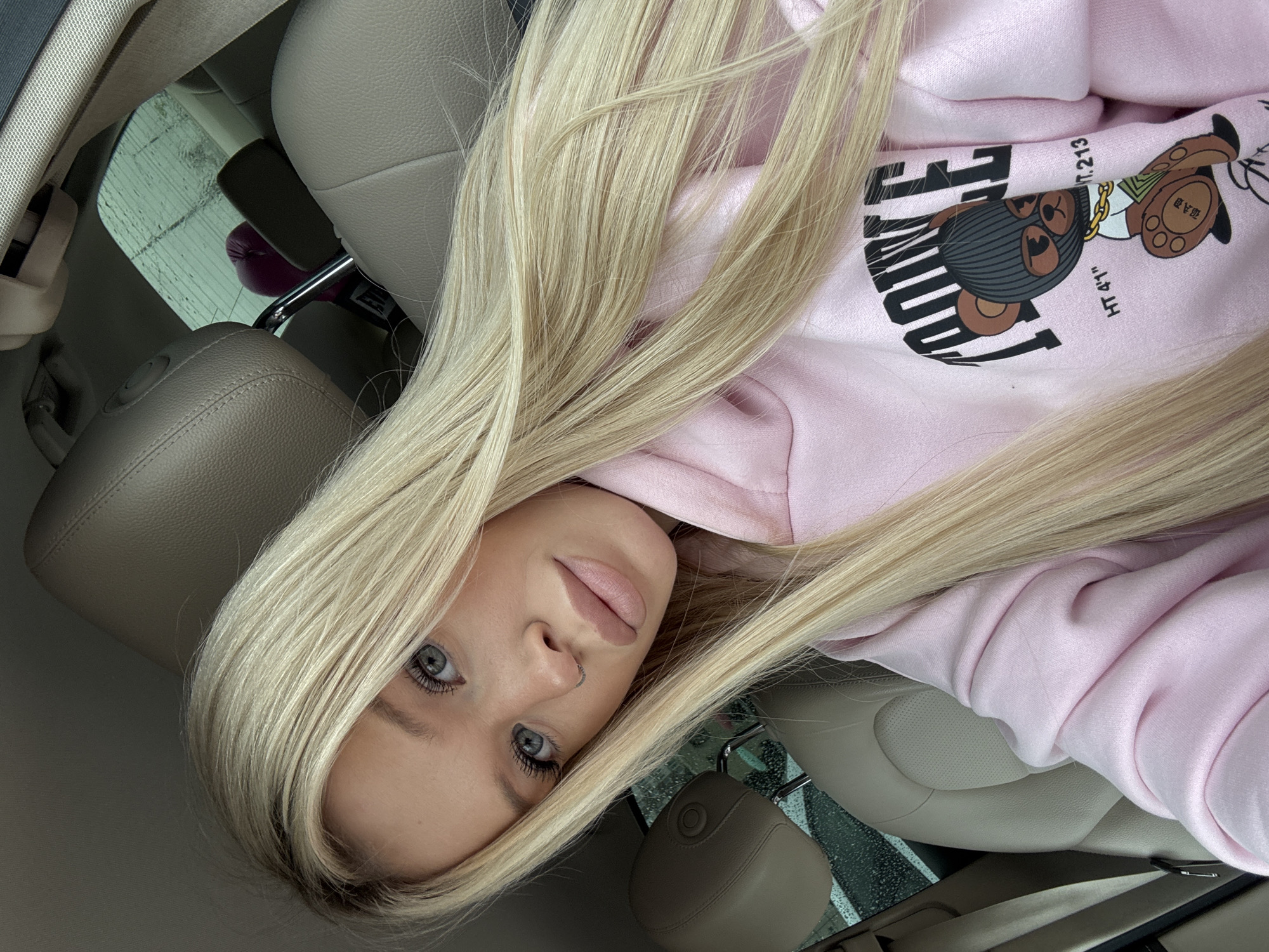 This is a portrait of a young Caucasian woman with long blonde hair and striking blue eyes. She is wearing a light pink hoodie with a graphic print of a cartoon bear wearing a ski mask and gold chains. The text "POWE" is visible on the hoodie, along with "HT.41" and "t. 213". She has on a small silver nose ring. The woman appears to be inside a car, as suggested by the beige leather car seats and seatbelt visible in the background. Her hair is styled loosely, with strands falling across her face and shoulders. The lighting suggests it might be daytime, possibly overcast, as the mood is somewhat subdued. There are indications of rain on the car's window. The scene is a close-up of the woman's face and upper body, with the background out of focus.
