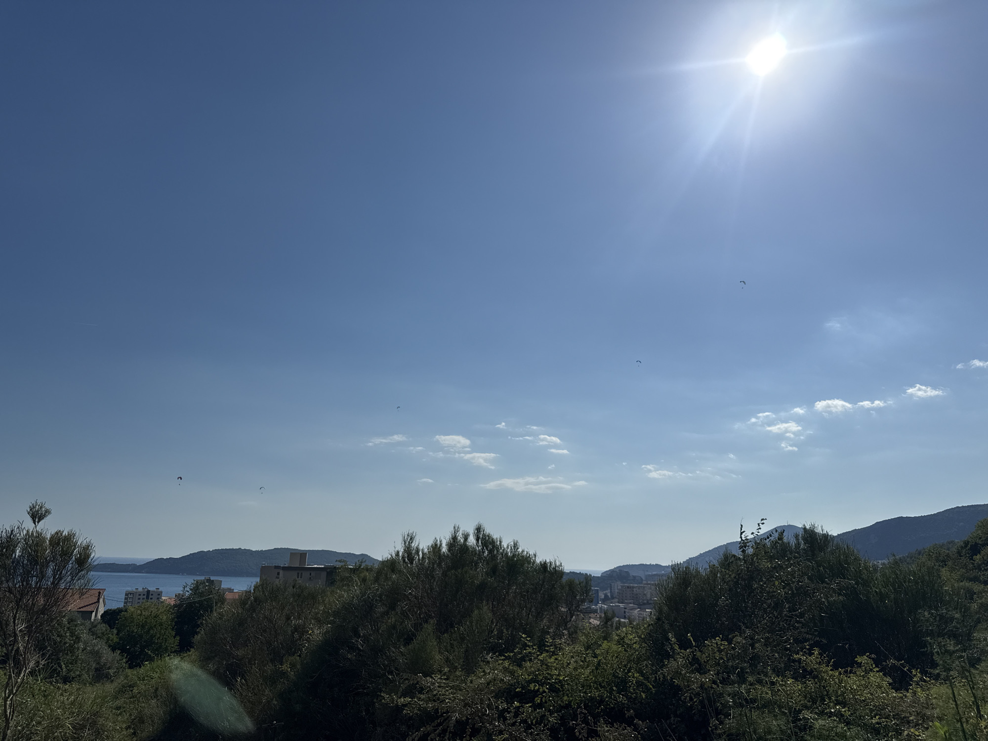 This media file depicts a scenic outdoor view, likely from an elevated vantage point, overlooking a coastal town and the sea. The bright sun in the upper right corner indicates it is daytime, possibly late morning or early afternoon, with clear skies. Several paragliders are visible in the sky, suggesting recreational activity.
The foreground is dominated by lush green foliage and trees, with some bare branches visible on the left. In the middle ground, a body of water, presumably the sea, stretches towards a distant coastline with rolling hills. A few buildings, including what appear to be apartment complexes or hotels, are nestled along the coast and further inland. The architecture suggests a Mediterranean or coastal European setting.
The overall mood is peaceful and serene, with the vast expanse of the blue sky and the calm water contributing to a sense of openness and tranquility. The paragliders add a dynamic element to the scene, hinting at adventure and enjoyment of the outdoors. No text is visible in the image. The location context places this scene in Rafailovići, Montenegro, which aligns with the visual cues of a coastal town with hilly terrain.
