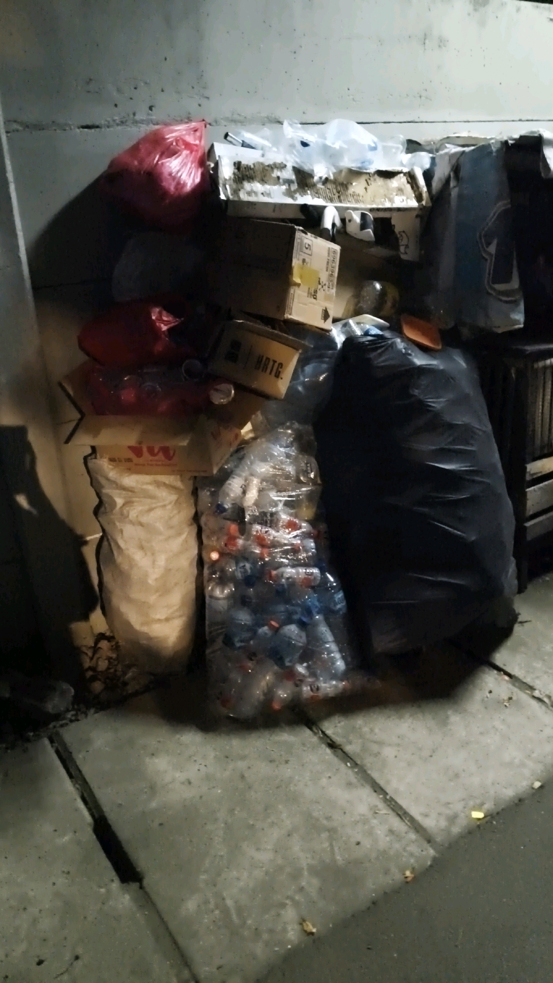 This image depicts a collection of refuse piled against a concrete wall. The refuse includes several plastic bags, some appearing to contain trash, and a large black garbage bag. There are also cardboard boxes, some of which are crushed or open, revealing their contents. A clear plastic bag filled with empty plastic bottles is visible, as is a large white woven bag. The ground is paved with concrete slabs, and debris is scattered on the surface. The scene appears to be an outdoor urban environment, possibly an alleyway or a public space. The lighting suggests it is nighttime or a dimly lit area. Without further information, it is impossible to confirm the specific location within South Jakarta, Indonesia.