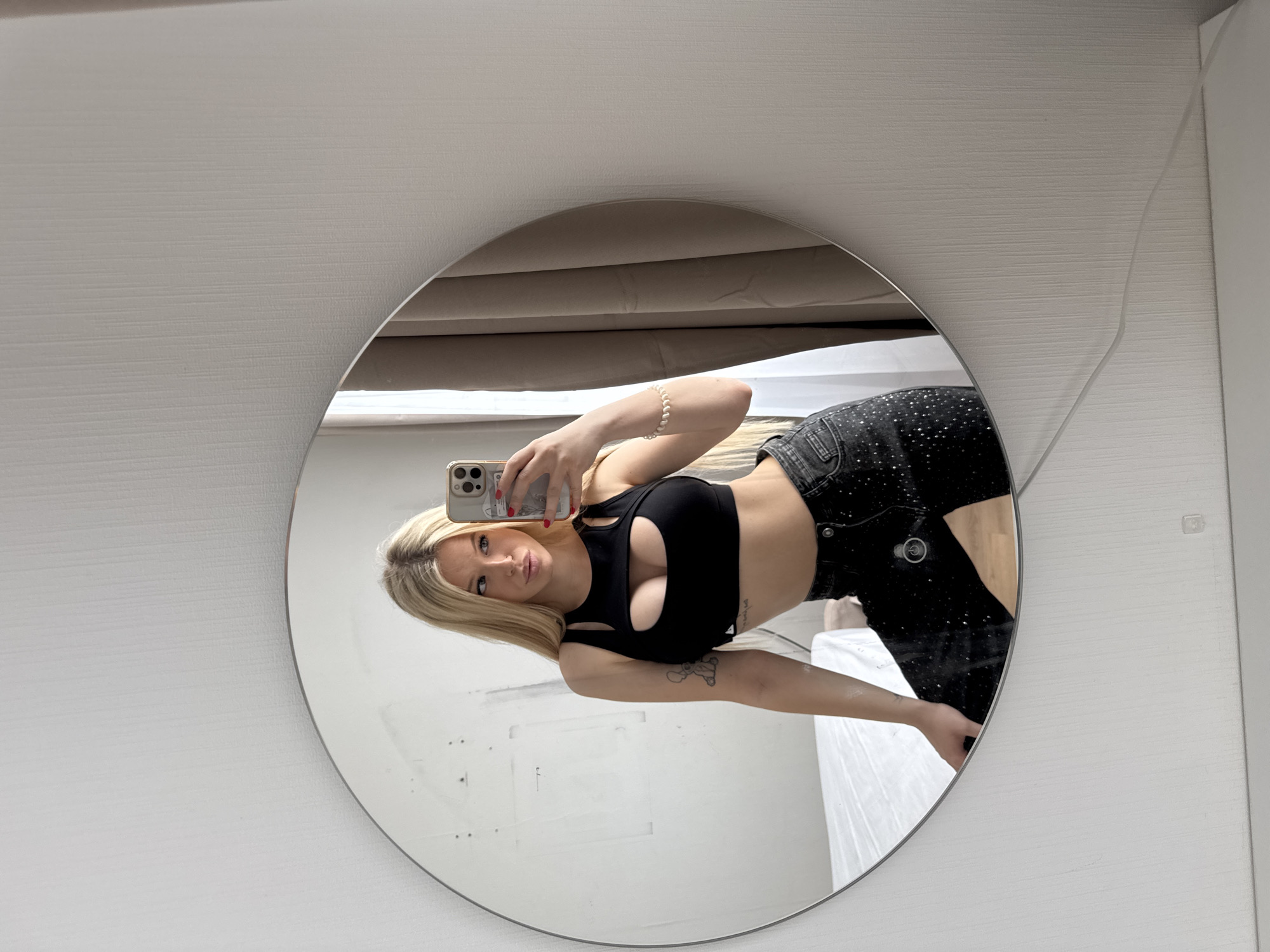 A young woman with blonde hair is taking a mirror selfie in a room. She is wearing a black crop top and black jeans with rhinestones. She is holding a smartphone in her right hand and her left arm is resting on her leg. The mirror reflects her body and a portion of the room, which appears to be a bedroom with a bed and curtains. There is no indication of Saint Petersburg, Russia in the image.