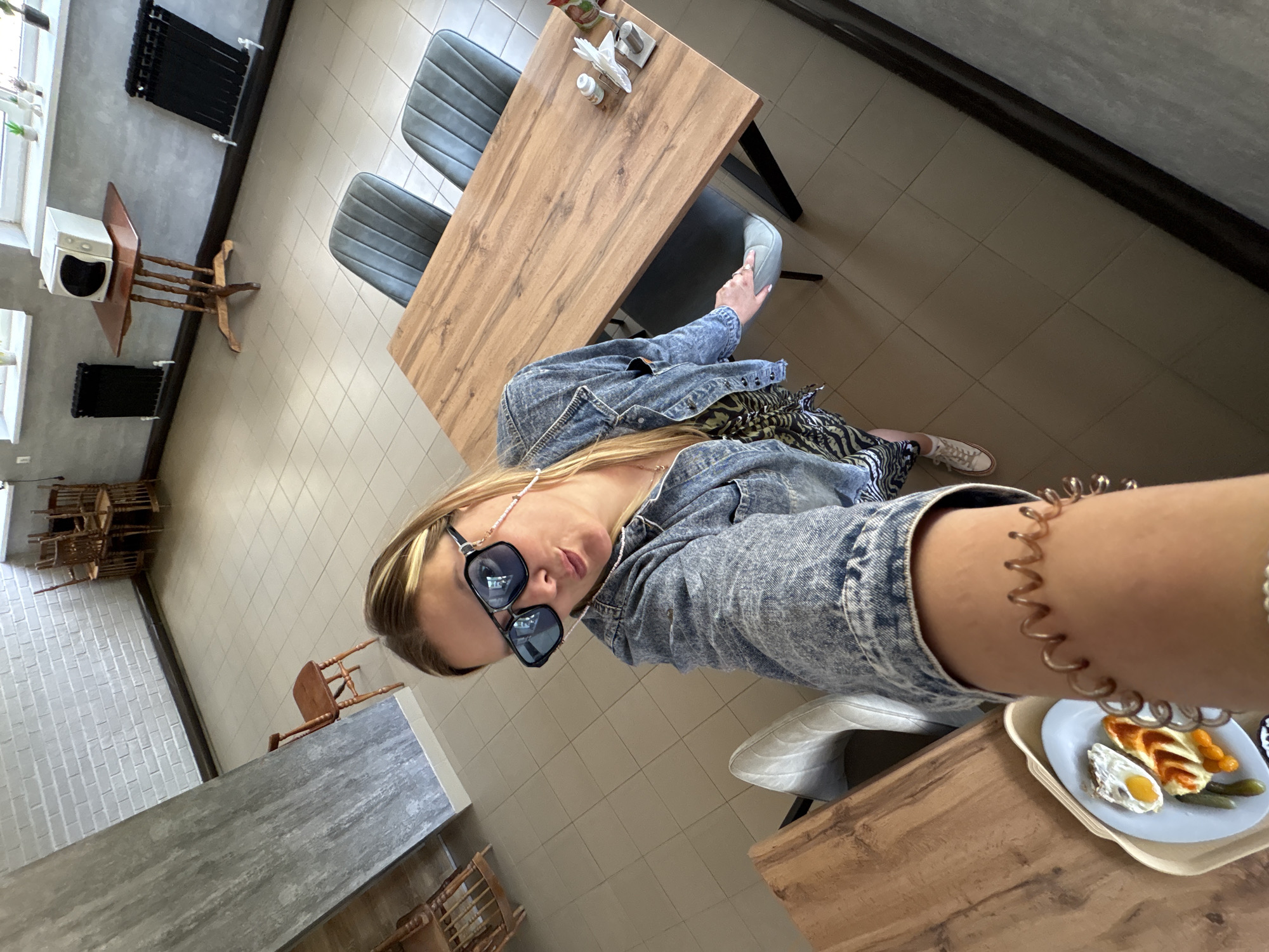 A young Caucasian woman with blonde hair and sunglasses is taking a selfie in what appears to be a cafe or dining area with a light-colored tiled floor. She is wearing a denim jacket and a patterned dress. In the foreground, a plate of food is visible, including what looks like a fried egg and some sort of pastry. Behind her, a wooden table with chairs is set up, and further in the background, there are more chairs stacked up. The walls are a light grey brick pattern. The overall impression is a casual dining setting.