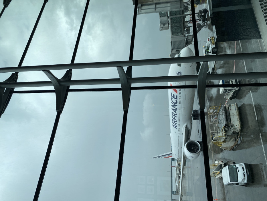 An Air France Boeing 777 aircraft is stationary on a wet tarmac at Charles de Gaulle Airport (CDG) in Mitry-Mory, France, viewed through large, multi-paned glass windows of an airport terminal. A grey passenger boarding bridge is connected to the aircraft's forward left door. Ground support equipment, including a white cargo loader and a white utility van, are positioned on the wet apron. The sky is heavily overcast, indicating dim, cloudy conditions, and the tarmac shows signs of recent or ongoing rain with visible puddles and reflections. No individuals are visible in the frame. The window structure consists of prominent grey vertical and horizontal beams with integrated supports.