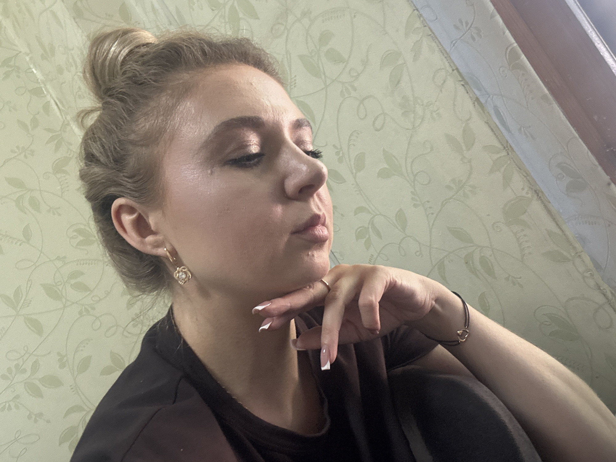 A young woman with blonde hair pulled into a bun is captured in a close-up, side-profile shot. She is wearing a dark top and rests her chin on her hand, her fingers adorned with French manicured nails and a gold ring. A delicate gold earring with a floral design is visible in her ear, and a thin black bracelet with a gold heart charm encircles her wrist. Her eyes are closed, and her makeup is subtly applied, highlighting her features. The background features a light green wallpaper with a faint floral pattern. The image is shot from a low angle, with the woman's face tilted downwards.