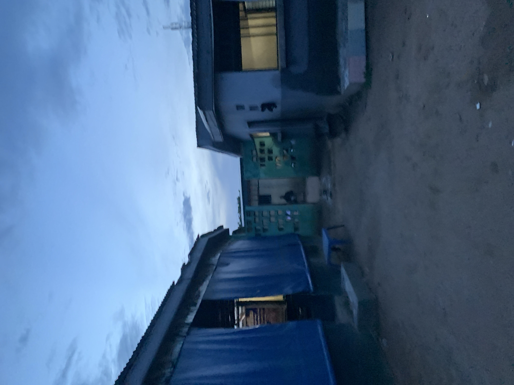 This image captures an outdoor scene at twilight in New Stadium, Nigeria. The lighting is low and evenly diffused, casting a blue-grey hue across the environment.
In the foreground to the left, a single-story structure is partially visible. Its roof is constructed of corrugated metal, and the front facade is obscured by two large, vertically hanging blue tarpaulins or fabrics. Interior light emanates from behind these coverings. A small, blue plastic stool rests on the unpaved ground directly in front of this structure.
To the right, another single-story building is present, featuring light-colored walls (possibly grey or light blue) and a corrugated metal roof. A rectangular window on its front face displays horizontal grilles or louvers, and interior illumination is visible. Indistinct features are present on its exterior wall. A tall, slender mast or antenna structure is faintly visible against the sky in the upper right background, above this building.
Centrally, in the background, a green metal gate or barrier with a grid-like or cut-out design connects the two structures or forms part of an adjacent property. A dark, indistinct human silhouette is observable near or behind this green gate.
The ground between and in front of the structures is unpaved, appearing to be bare earth or dirt, scattered with small, light-colored debris. The sky above is a pale blue, transitioning to darker grey with diffused cloud formations.