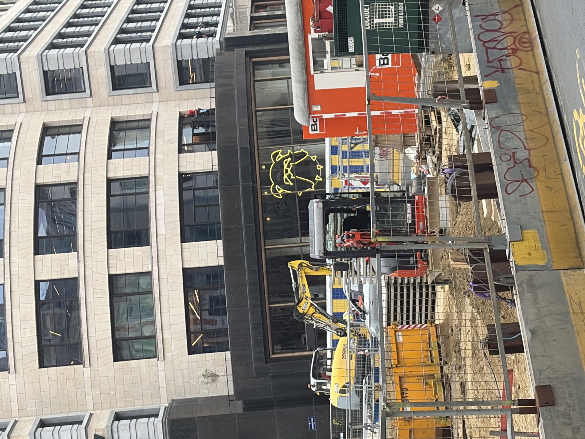 This image captures a construction site in the city of Brussels, Belgium. In the foreground, a wire mesh fence encloses the area, revealing construction materials and equipment. A yellow skip is visible to the left, and stacks of grey concrete slabs are present in the middle ground. A yellow excavator with a black cab is positioned on the left, and a smaller red and black excavator is on the right. Behind the construction equipment, a bright orange portable building with a white door and windows is situated. To the left of the orange building, a large, yellow neon outline of a stylized dog's head is visible on a dark facade. In the background, a multi-story building with a beige, stone-clad facade and dark-framed windows rises above the construction site. A few individuals are visible on a balcony or walkway of this building, one wearing a red shirt and another in dark clothing. The environment appears to be an urban setting, indicated by the presence of the large building and the construction activity.