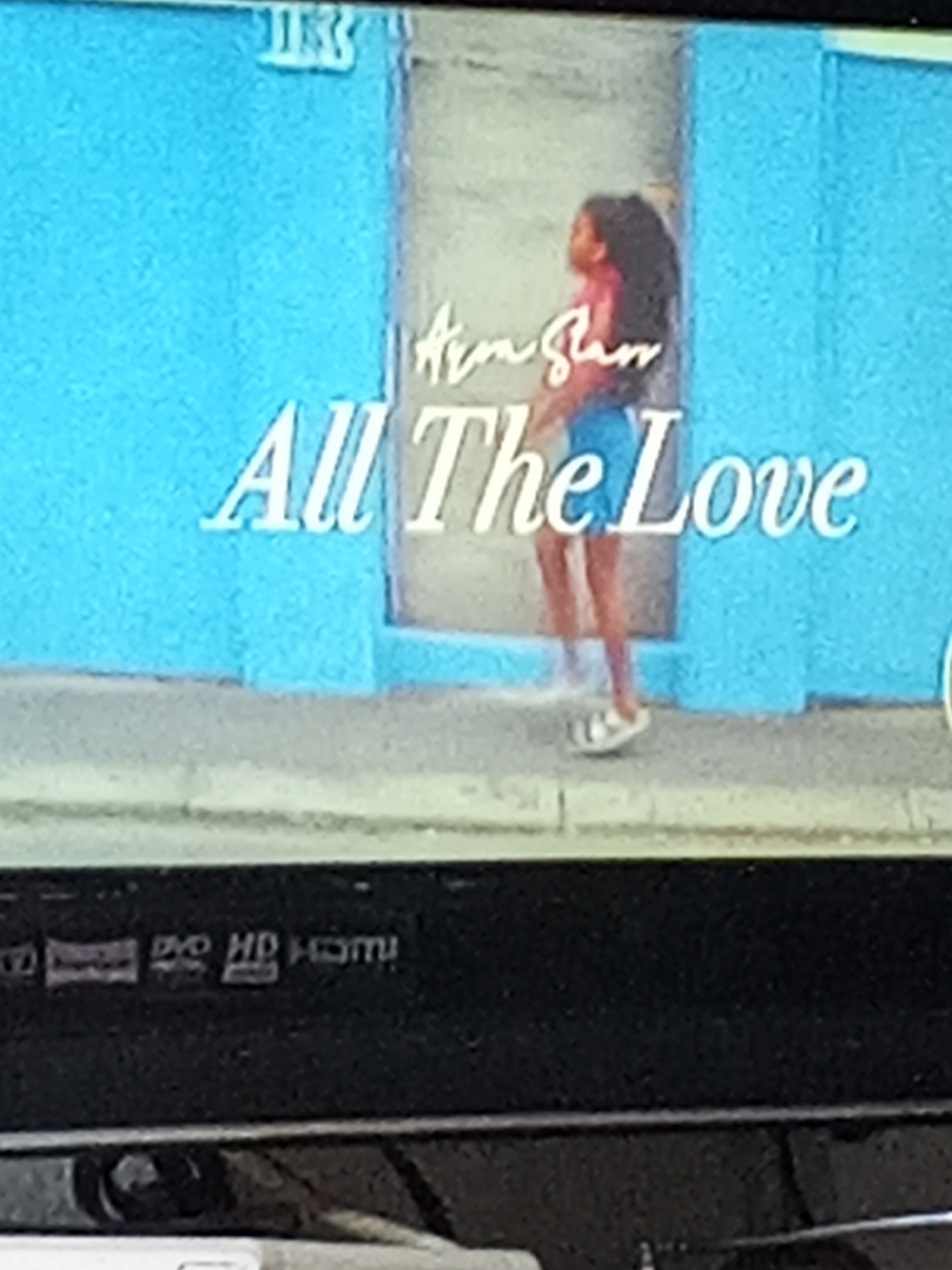 A still image shows a young woman in a blue outfit, possibly a dress or a top and shorts, with her back to the viewer. She stands on a sidewalk in front of a light blue building in Gwong, Nigeria. The wall has the text "Airm Slaw" and "All The Love" written in white. The video is showing on a screen which features the labeling "DVD, HD, HDMI" at the bottom of the frame.