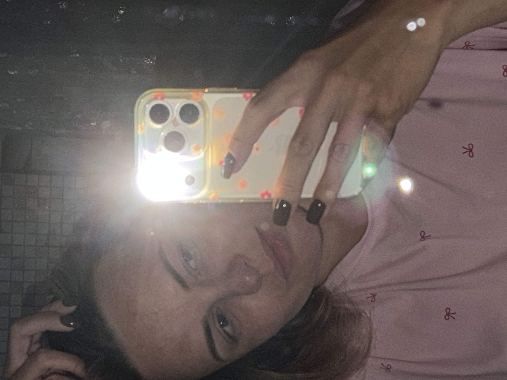 A woman is taking a mirror selfie in what appears to be a bathroom in Votkinsk, Russia. She is wearing a pink shirt with a repeating pattern of red bows. Her right hand is holding a smartphone with a transparent case adorned with small floral designs. The phone is angled to capture her reflection. She is looking at her reflection in the mirror, which shows her face and upper body. The background of the mirror appears to be a tiled surface, possibly a shower wall, with some water spots or smudges. There is a bright light source reflecting in the mirror, likely from a flash or overhead lighting, which causes some glare and lens flare across the image. Her hair is dark and she has dark nail polish on her fingers.