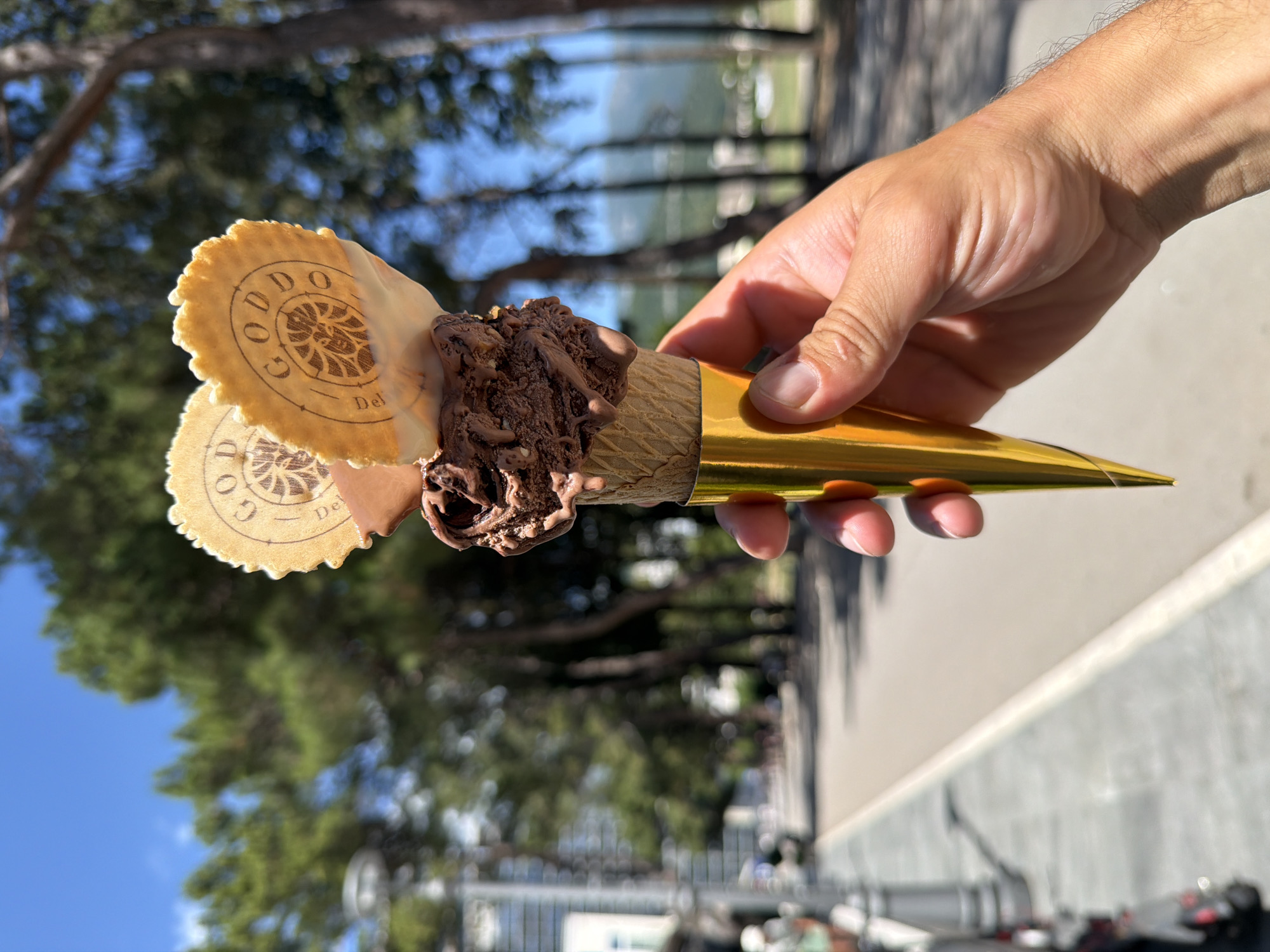 A person's right hand, with tanned skin, holds an ice cream cone centrally in the foreground. The cone contains two scoops of dark chocolate ice cream, exhibiting visible melt, chocolate drizzles, and embedded fragments. Two round, serrated-edge wafers, each embossed with the text "GODDO" and "Del" surrounding a stylized central emblem, are partially inserted into the top scoop. White frosting or glaze cascades from the wafers onto the ice cream. The lower portion of the waffle cone is enveloped in a conical, reflective gold-colored wrapper. The background depicts an outdoor urban environment under a clear blue sky on a brightly lit day. A paved road or pathway extends from the midground, bordered on the right by a dense line of tall, dark green coniferous trees. On the left, a grey sidewalk features a partial view of a lamppost and a parked scooter, leading to blurred distant buildings. Faint outlines of mountains are discernible on the far right horizon. This scene is situated in the city of Budva, Montenegro.