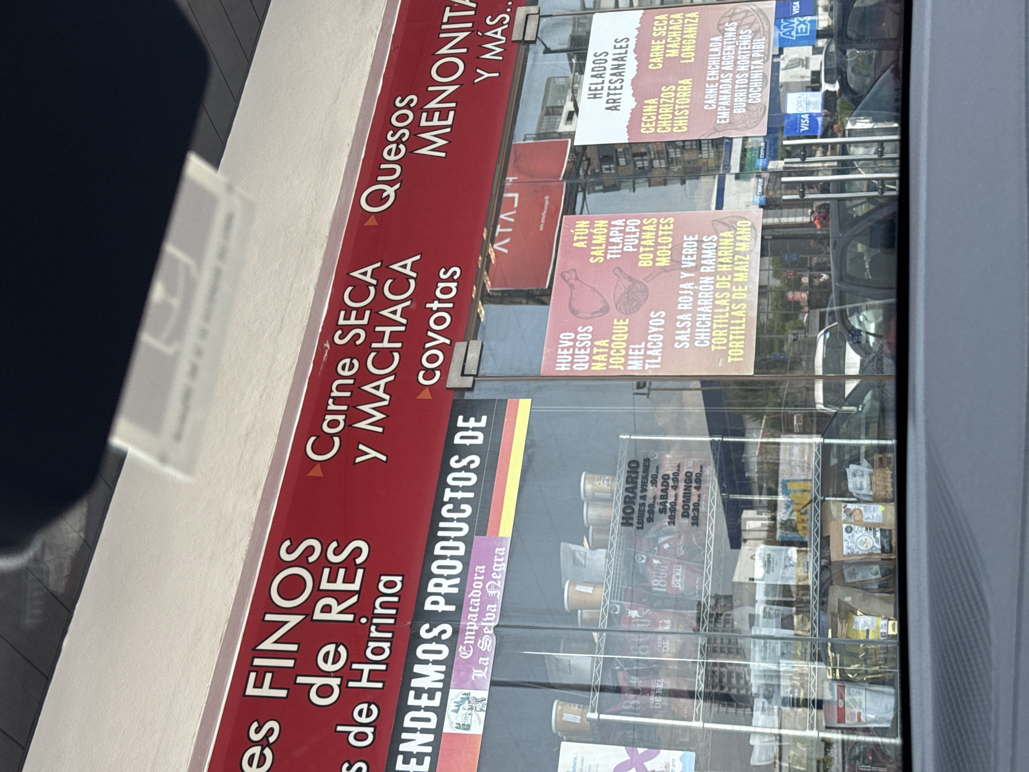 Here is a description of the image:
The image shows the storefront of a business in Heroica Puebla de Zaragoza, Mexico, that sells a variety of food products. The storefront has large glass windows with various signs advertising the products sold inside.
The sign along the top of the storefront reads "es FINOS de RES y MACHACA coyotas Quesos MENONITA Y MÁS..", indicating they sell fine beef and Machaca products, along with Mennonite cheeses. Another sign advertises "ENDEMOS PRODUCTOS DE" and includes the logo for "Empacadora La Selva Negra."
Additional signs visible through the glass list other products such as artisan ice cream, meats like cecina, chorizo, chistorra, and longaniza, and dishes such as carne enchilada, empanadas argentinas, burritos norteños, and cochinita pibil. Another sign lists ingredients like eggs, cheese, cream, jocoque, honey, tilapia, octopus, and molotes. The store also sells tortillas.
The business hours are displayed: Monday to Friday 9:00 - 7:00, Saturday 10:00 - 4:00, and Sunday 10:30 - 4:00.
Inside the store, shelves stocked with packaged food products are visible. Payment options include Visa and American Express. Reflections in the glass windows show cars, buildings, and the street environment outside the store.