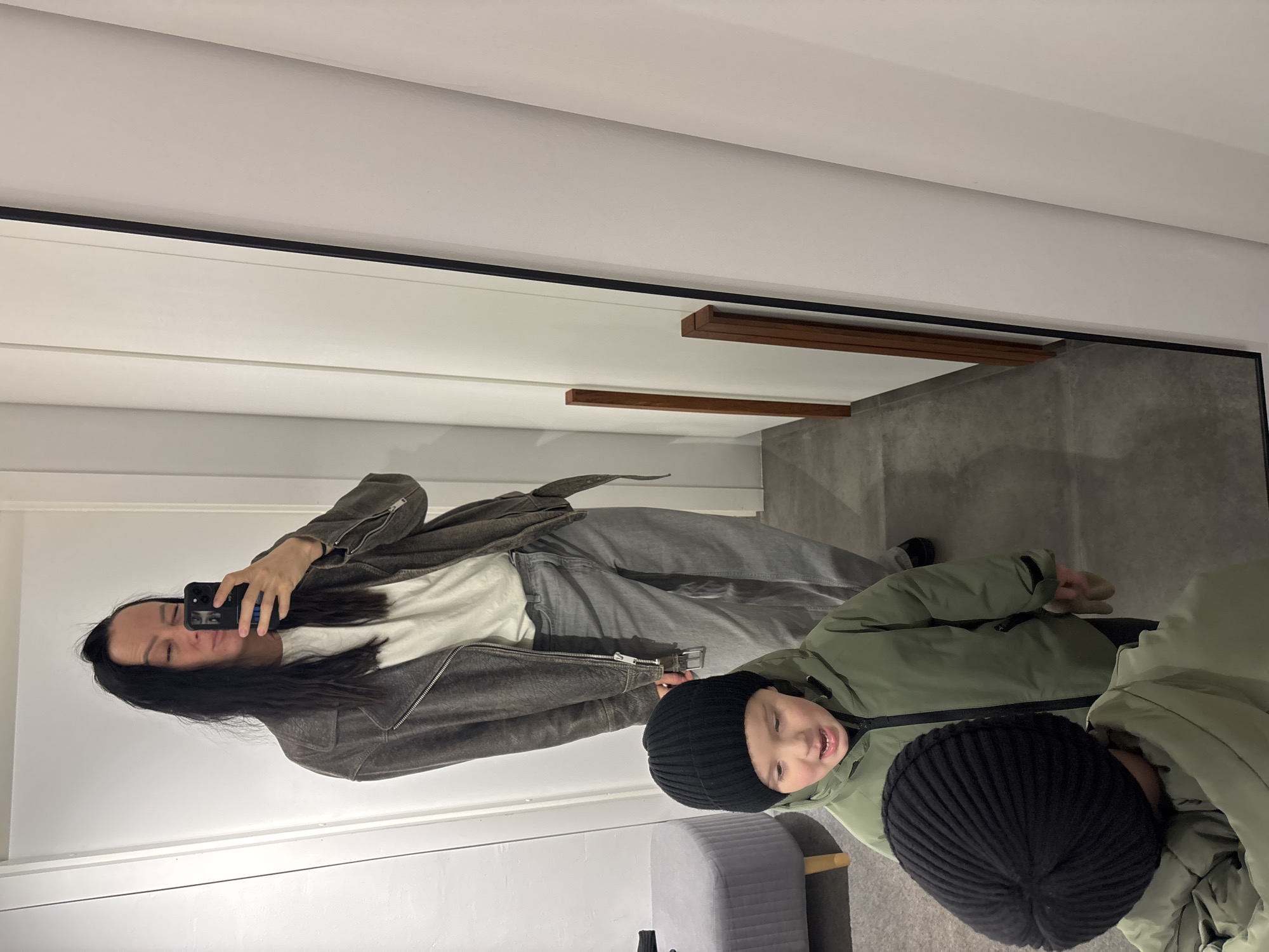 A woman is taking a selfie in a full-length mirror, with a child also visible in the reflection. The woman is wearing a brown jacket, a white t-shirt, and grey jeans. She is holding a black smartphone in her right hand. The child, wearing a green jacket and a black knit hat, is partially visible next to her. The environment appears to be an interior space with a concrete floor. The background includes white walls and a grey upholstered stool or bench. The location in Chaikovsky, Russia, cannot be determined from the image itself.