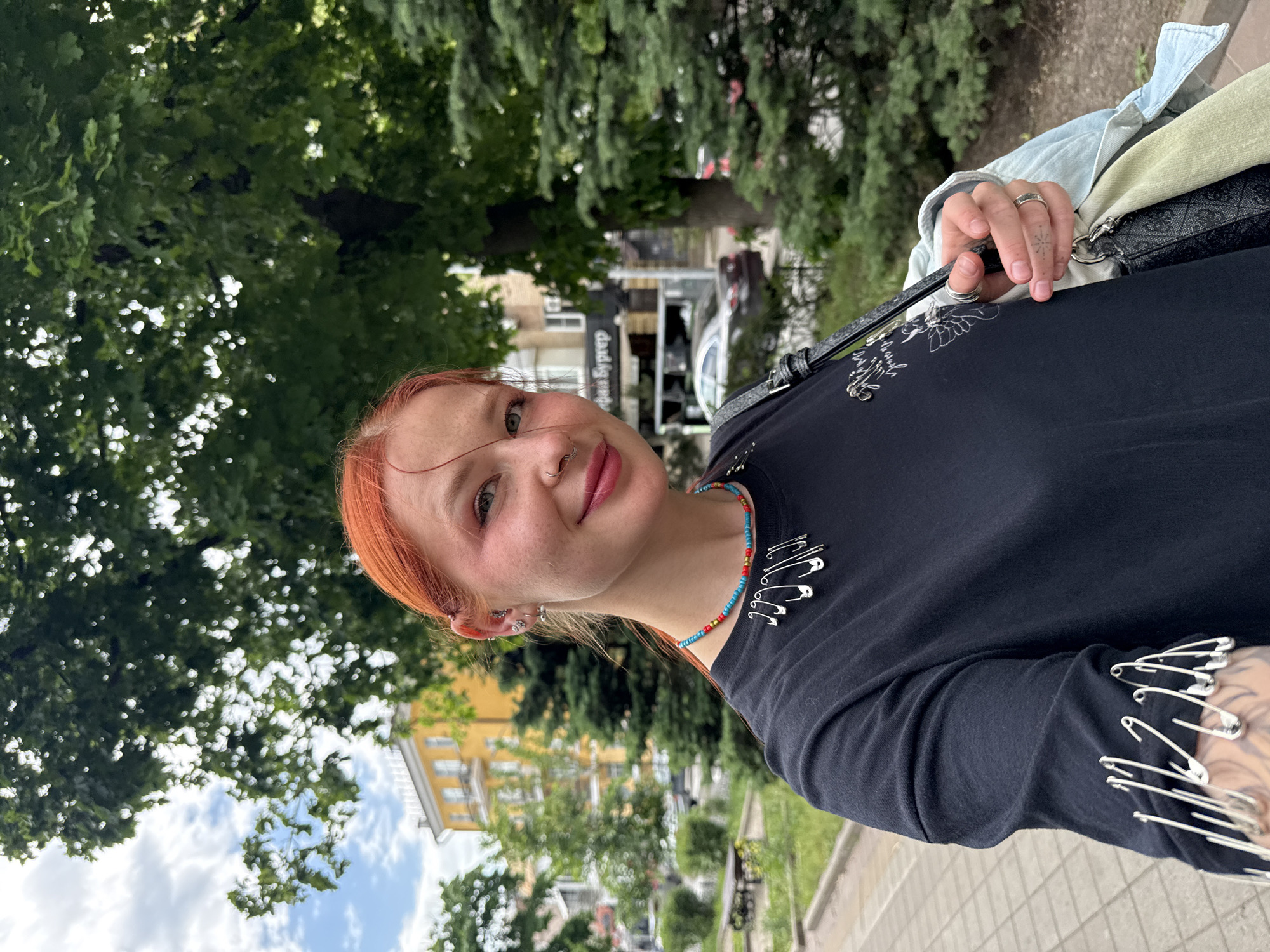 The image depicts a young woman with red hair and fair skin, posed outdoors in a seemingly urban environment. She is wearing a black long-sleeved shirt adorned with safety pins and a beaded necklace. A handbag is slung over her shoulder. The background shows trees, a street, a building with the word "prepay" or "Syprep" on its facade, and several parked cars. Based on the building style and the presence of Russian text, the location is likely in a city in Russia. Given the provided context, the location is identified as Rostov-on-Don, Russia. The image appears to be a casual snapshot or selfie.
