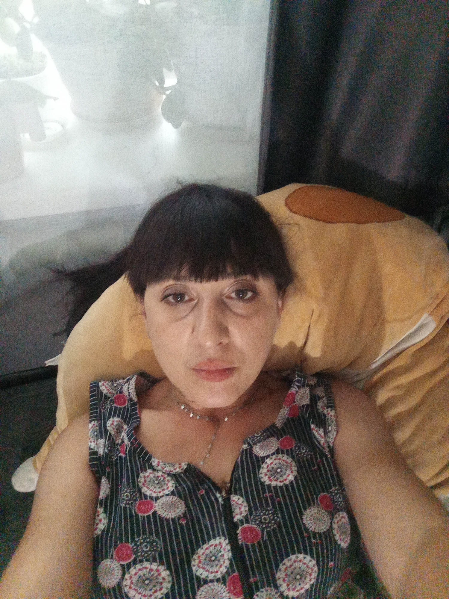 A woman with dark hair and bangs, wearing a sleeveless dress with a circular pattern, lies on a large, light brown pillow. She is looking directly at the camera. The background is blurred and appears to be a window with plants visible, and a dark curtain. The image was taken in the city of Belogorsk, Russia.
