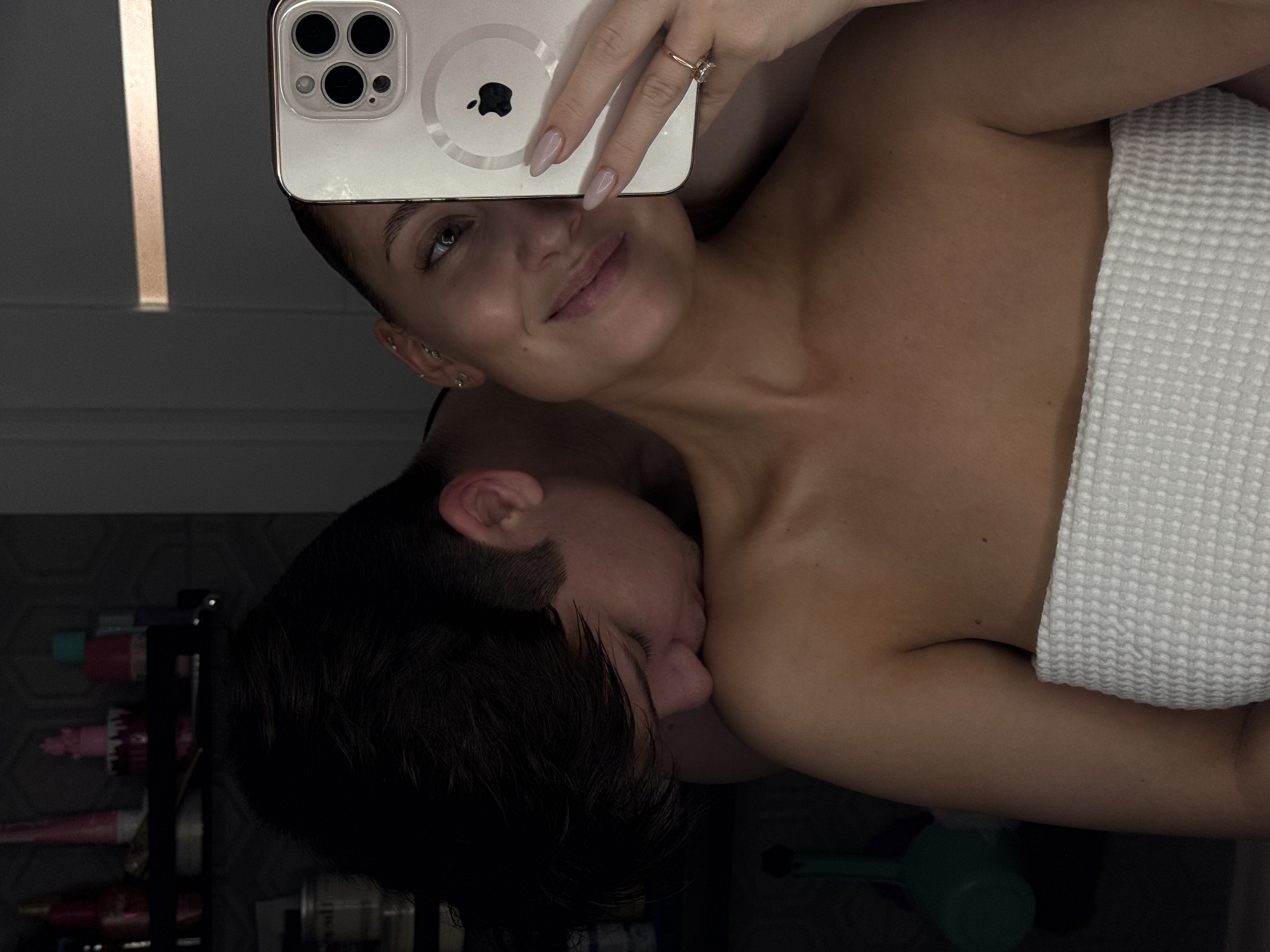 A mirror selfie captured in a dimly lit indoor setting, likely a bathroom, features a woman and a man. The woman, foregrounded, wears a white waffle-knit towel wrapped around her chest and smiles faintly towards the camera. She holds a light-colored iPhone with a clear case in her left hand, which displays light-colored manicured nails and a ring with a clear stone on the ring finger. Behind her, a man with dark hair is seen with his face pressed against her left shoulder, appearing to kiss or nuzzle it. The background consists of a dark wall with a subtle geometric pattern, a dark shelf stocked with various toiletries including pink and teal bottles, and a teal plastic ladle hanging on the left. A reflective surface or light fixture is partially visible on the right. The specific location within Chaikovsky, Russia, cannot be definitively identified from the visual evidence presented.