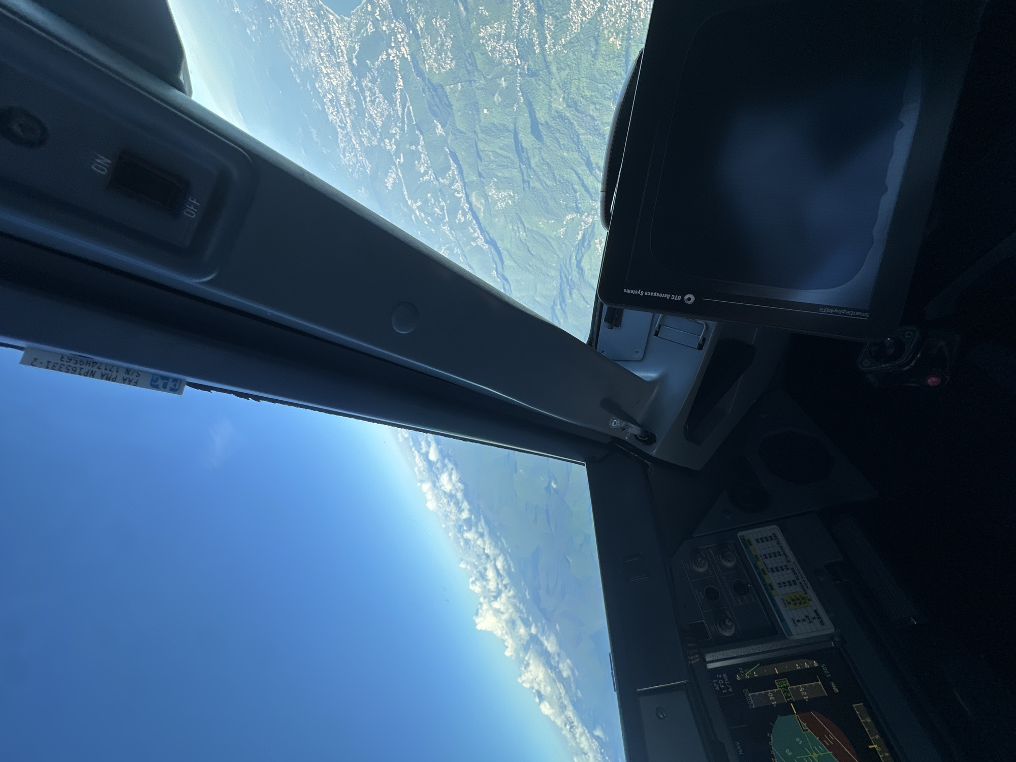 The image appears to be taken from the cockpit of an aircraft in flight. The view outside the aircraft shows the Earth's surface, with a mix of green terrain and what appears to be some cloud cover near the horizon. Part of the cockpit is visible, including various instruments, screens, and controls.
Based on the OCR text, some of the displayed information includes navigation data (NAV), autopilot settings (AP1, FD 2, A/THR), altitude (20000), vertical speed (+1.30-), airspeed (125), and a QNH value (1005). The term "SmartDisplay@EFB" and "UTC Aerospace Systems" are also visible, likely referring to the electronic flight bag system. A switch with "ON" and "OFF" positions is also visible.
There is no indication in the image or OCR text that the location is in the city of Khimki, Russia.