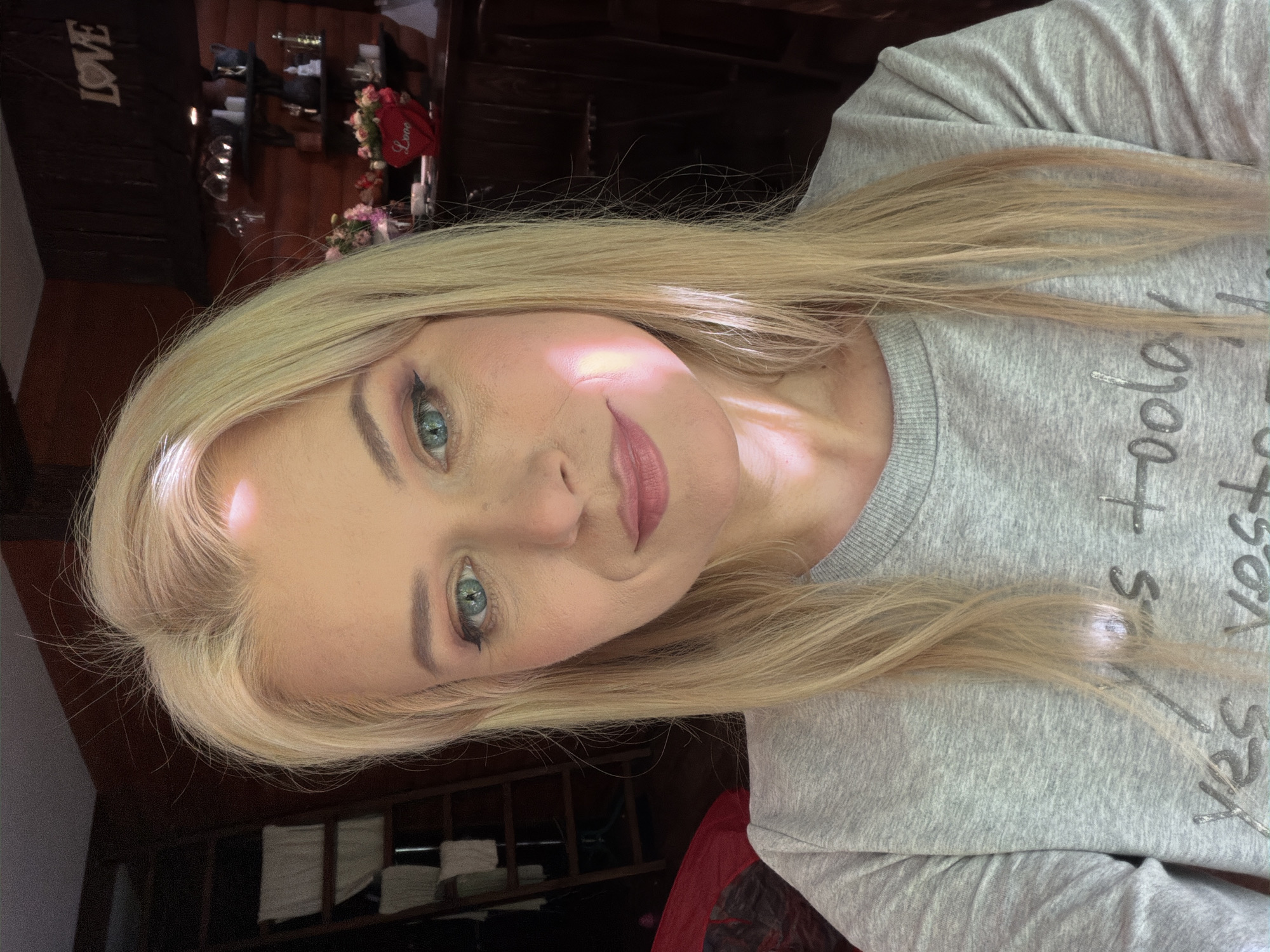 A blonde woman with blue eyes and light makeup is taking a selfie in a rustic interior setting. She is wearing a gray sweatshirt with text that appears to say "yes today" and "yes yesterday." The background features wooden walls, shelves with decorative items including flowers and a red heart with the word "Love," and a ladder with folded white towels. A beam of light illuminates the woman's face from the right side. The location is identified as Sochi, Russia.