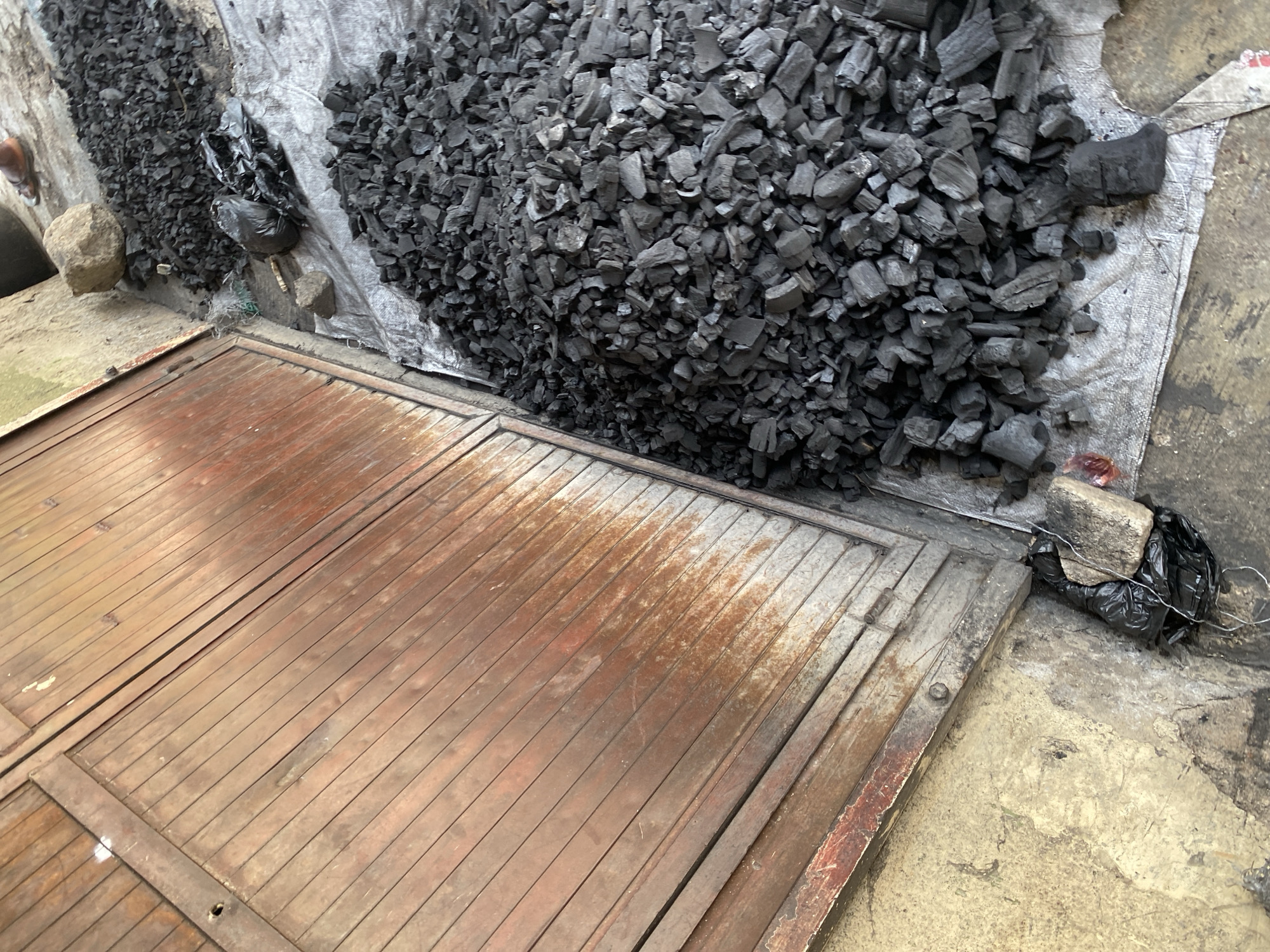 The image shows a pile of charcoal on a white tarp in front of a weathered wooden gate in the city of Jos, Nigeria. The charcoal is broken into irregular pieces, ranging in size from small fragments to larger chunks. Some pieces appear to have a rough, porous texture. The wooden gate is made of vertical planks, with signs of rust and wear, suggesting it is old and has been exposed to the elements. The ground around the tarp is a rough, concrete-like surface, with some dirt and debris visible. In the background, a small pile of charcoal is also visible, along with a black plastic bag and two rocks. The overall scene suggests a market or storage area where charcoal is being sold or prepared.