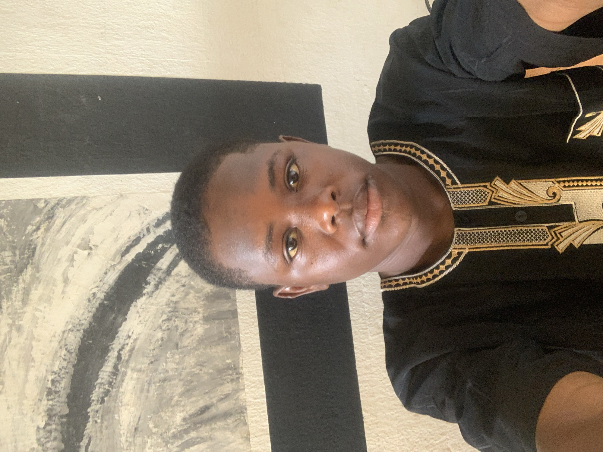 A portrait, rotated 90 degrees clockwise, features a young person with dark skin and short, closely cropped dark hair, looking directly at the camera. Their eyes are light brown or amber. The individual is wearing a black garment adorned with intricate gold and light-colored embroidery around the neckline, shoulders, and down the front, suggestive of traditional Nigerian attire.
The background shows an interior wall, divided horizontally into an upper section of light-colored paint and a lower section of black paint. To the lower left, an abstract piece of art or a textured wall section is visible, featuring bold brushstrokes in white, gray, and black. The lighting is bright and even, indicating an indoor setting. The person's expression is neutral and direct. No specific activity is taking place other than posing for the photograph. The scene depicts a personal portrait, likely taken in a contemporary indoor environment in Jos, Nigeria. There is no visible text in the image.