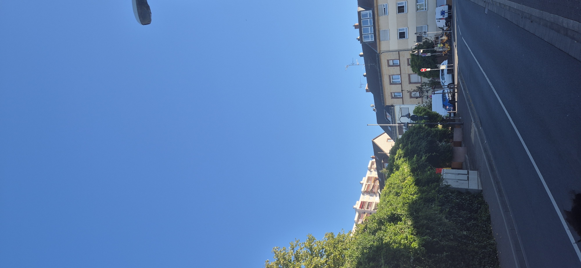 The image depicts a street scene in Mainz, Germany, under a clear blue sky. On the left, a dense green hedge lines a sidewalk. Behind the hedge, a modern apartment building with red balconies is visible. The street itself has a white line marking the lanes.
Further down the street, several residential buildings with tiled roofs are situated. These buildings exhibit typical German architecture with multiple windows and some have aerials on the roofs. Among the buildings, there are streetlights and traffic signs, including a red and white circular sign that likely indicates a traffic regulation.
Several vehicles are present on the street, including a blue car, a white car, a yellow construction vehicle (possibly a small excavator or front loader), and a white van. A traffic light is visible near one of the buildings. The overall impression is a well-maintained, residential street in a German city.