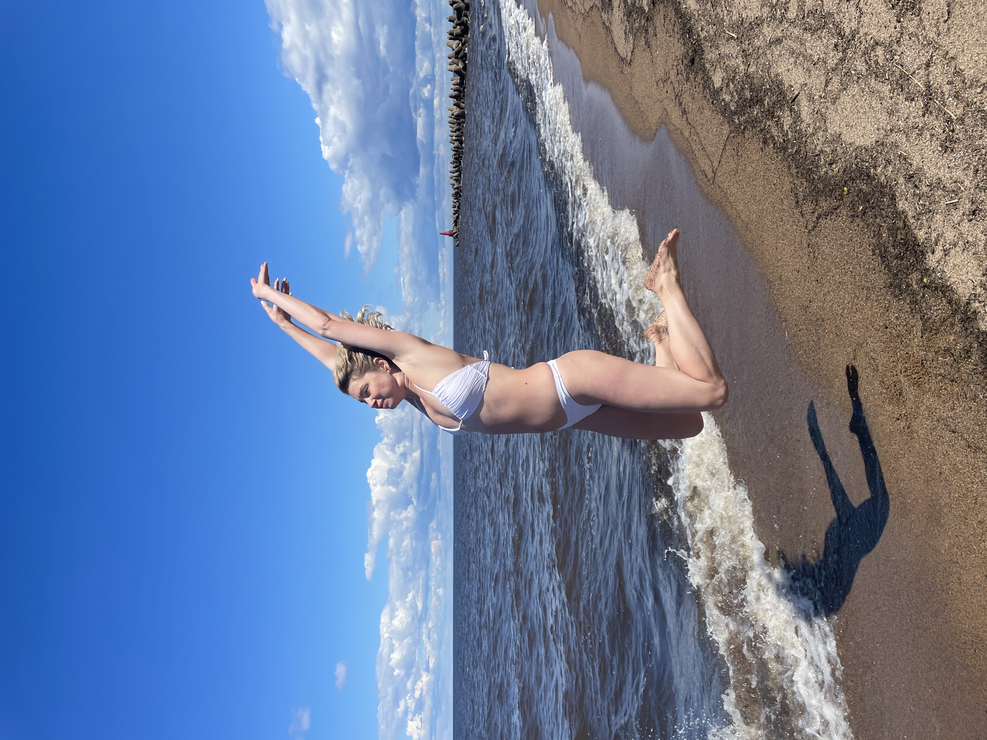 A woman in a white bikini is captured mid-air as she jumps on a sandy beach with gentle waves lapping at the shore. The sea extends to the horizon, where a line of concrete breakwaters, punctuated by a red marker, is visible under a bright blue sky dotted with white clouds. The woman's arms are raised above her head, and her body is arched in a dynamic pose. Her shadow is cast on the wet sand near the water's edge. The location is the shore of Lake Beloye in Belozersk, Russia.