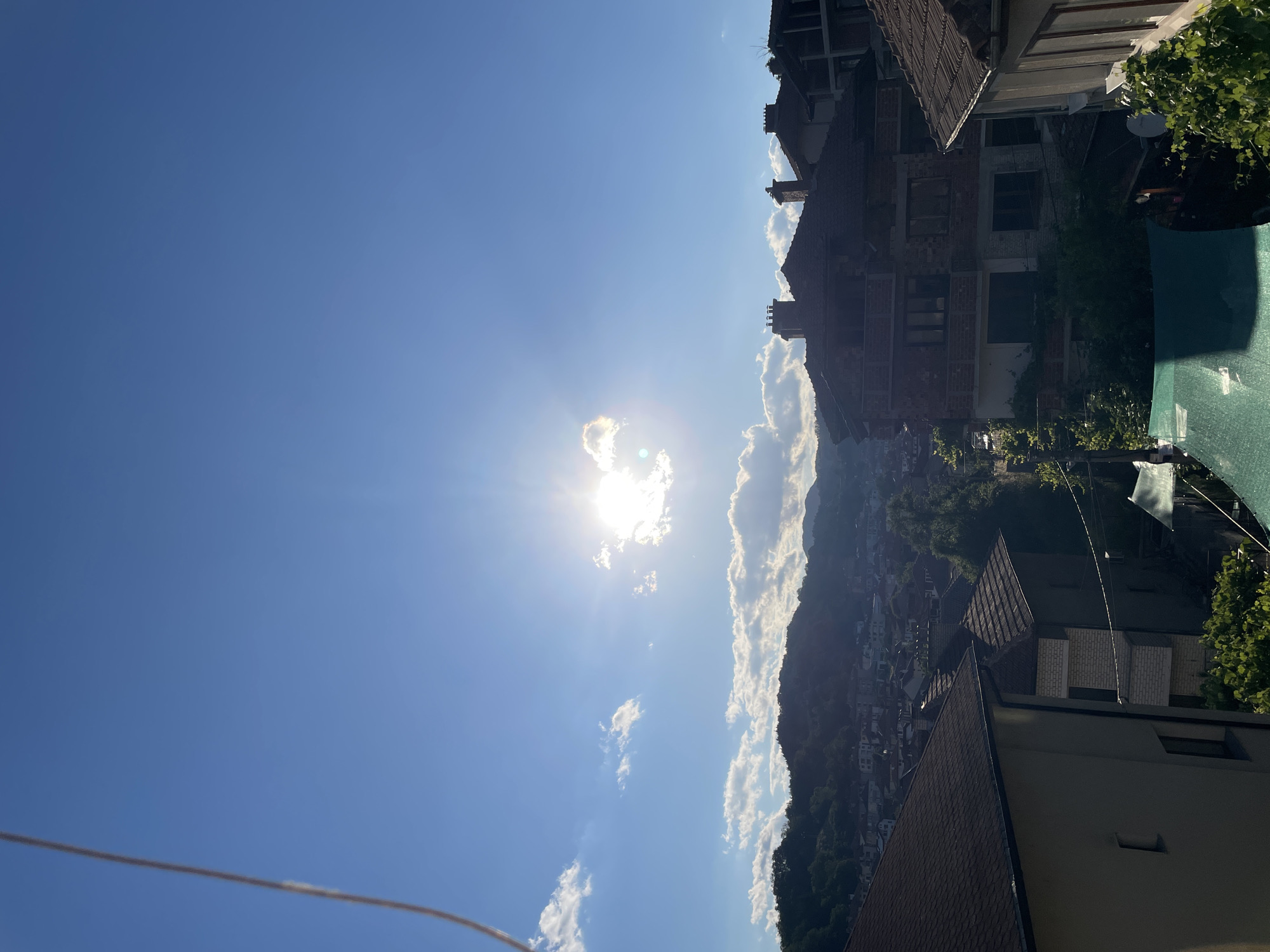 The image captures an elevated view of a residential area in Zlatograd, Bulgaria, under a bright, sunlit sky. The sun, a brilliant, white orb, is prominently positioned in the upper center, with rays of light emanating outward, partially obscured by a fluffy white cloud. The sky is a vibrant, clear blue, with scattered white clouds dotting the expanse.
In the foreground, the rooftops of several houses are visible, featuring dark, textured tiles. The architecture suggests a traditional Balkan style, with some houses appearing to be under construction or renovation, evident by exposed brickwork and incomplete structures. Chimneys are visible on some of the roofs.
Behind the immediate foreground, a hillside settlement unfolds, with numerous houses clustered together, their roofs a mosaic of terracotta and darker hues. Lush green trees and vegetation are interspersed throughout the residential area, softening the urban landscape.
The background is dominated by rolling hills covered in dense green foliage, indicating a mountainous or hilly terrain surrounding the town. The overall impression is one of a peaceful, sun-drenched afternoon in a Balkan town. There are no people or animals visible in the frame. The scene is static, with no apparent actions or interactions taking place. The environment is a mix of built structures and natural landscapes.