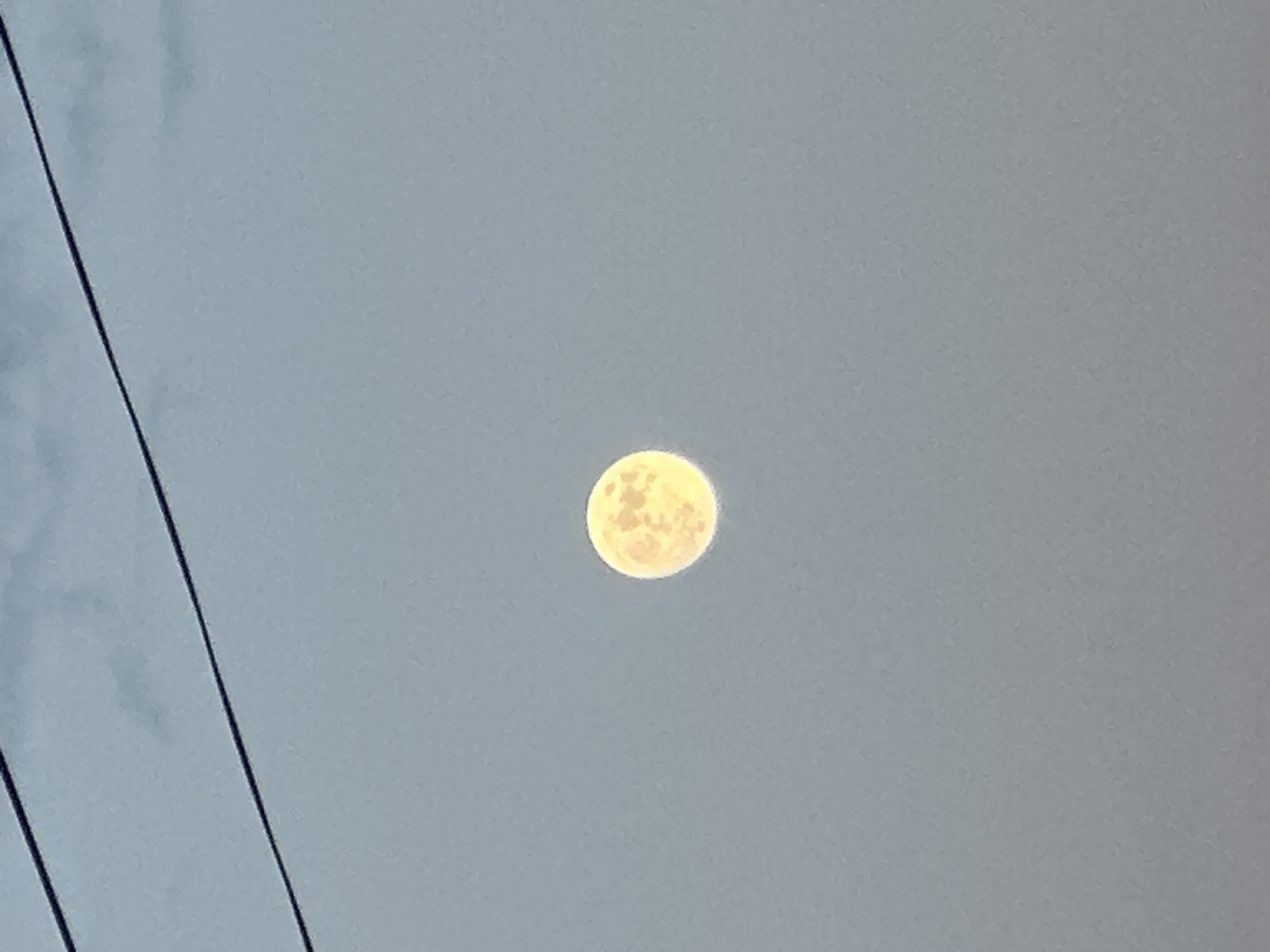 The image depicts a full moon in a gray sky. A pair of black power lines are visible in the left side of the frame. The location is not discernible.