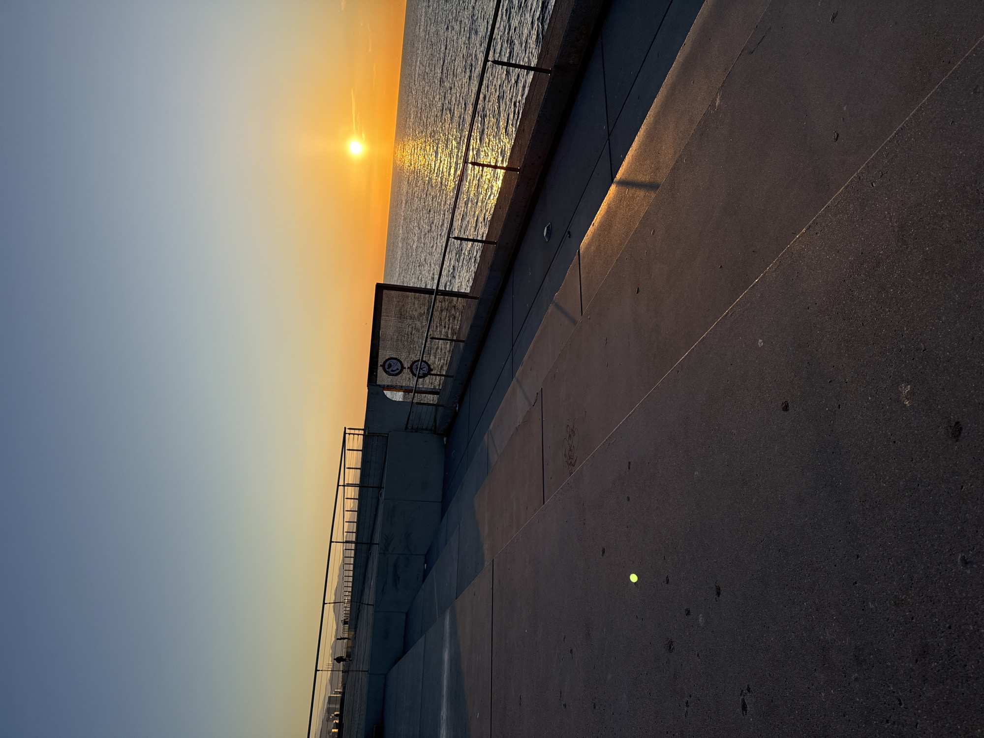 The image shows a sunset over the sea in Barcelona, Spain. The setting sun is reflected on the water. A concrete promenade with railings runs along the coast. The perspective is from the promenade looking toward the sea. The sky is a gradient from blue to orange.