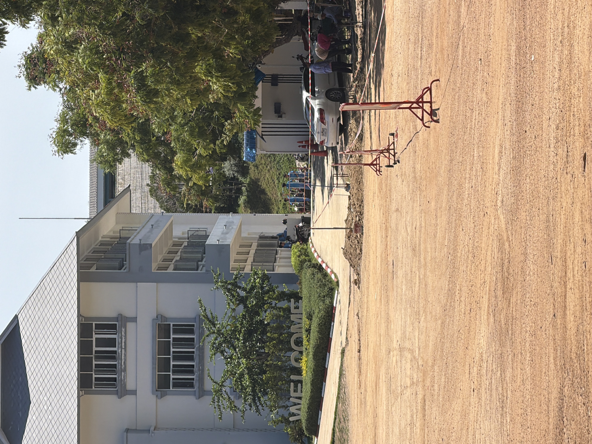 In Baan Bo Phut, Thailand, the scene depicts a construction site in front of a multi-story building. The building, painted in white and light gray, features a distinctive pitched roof with diamond-shaped tiles. In the foreground, a large area of bare, sandy earth is visible, with red metal barriers and caution tape sectioning off parts of the area. Several people, dressed in casual attire, are observed engaged in construction work, with some handling tools and materials. A white sedan is parked nearby, adjacent to a small white structure. A "WELCOME" sign, formed by large stone letters, is situated on a manicured hedge in front of the building, bordered by red and white striped curbing. Lush green trees and bushes surround the building and the sign, with a clear blue sky overhead.