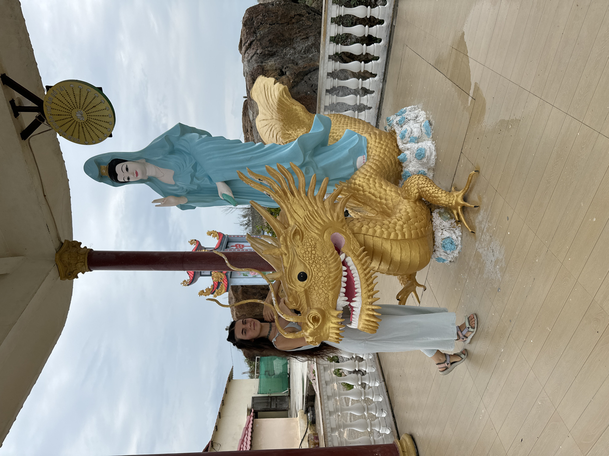 A young woman with long, dark hair is standing next to a golden dragon statue, positioned outdoors with light-colored tile flooring. The woman is wearing a light-colored dress and sandals. She is partially obscured by the head of the dragon. Behind the dragon is a statue of a blue-clad figure and a rocky outcrop. The woman is located at the Ong Dia Rock Cape temple (also known as the Mui Ne Dragon Temple) in Mui Ne, Vietnam. A building with a white railing and partial structures with red roofing are visible in the background, and the sky is overcast.