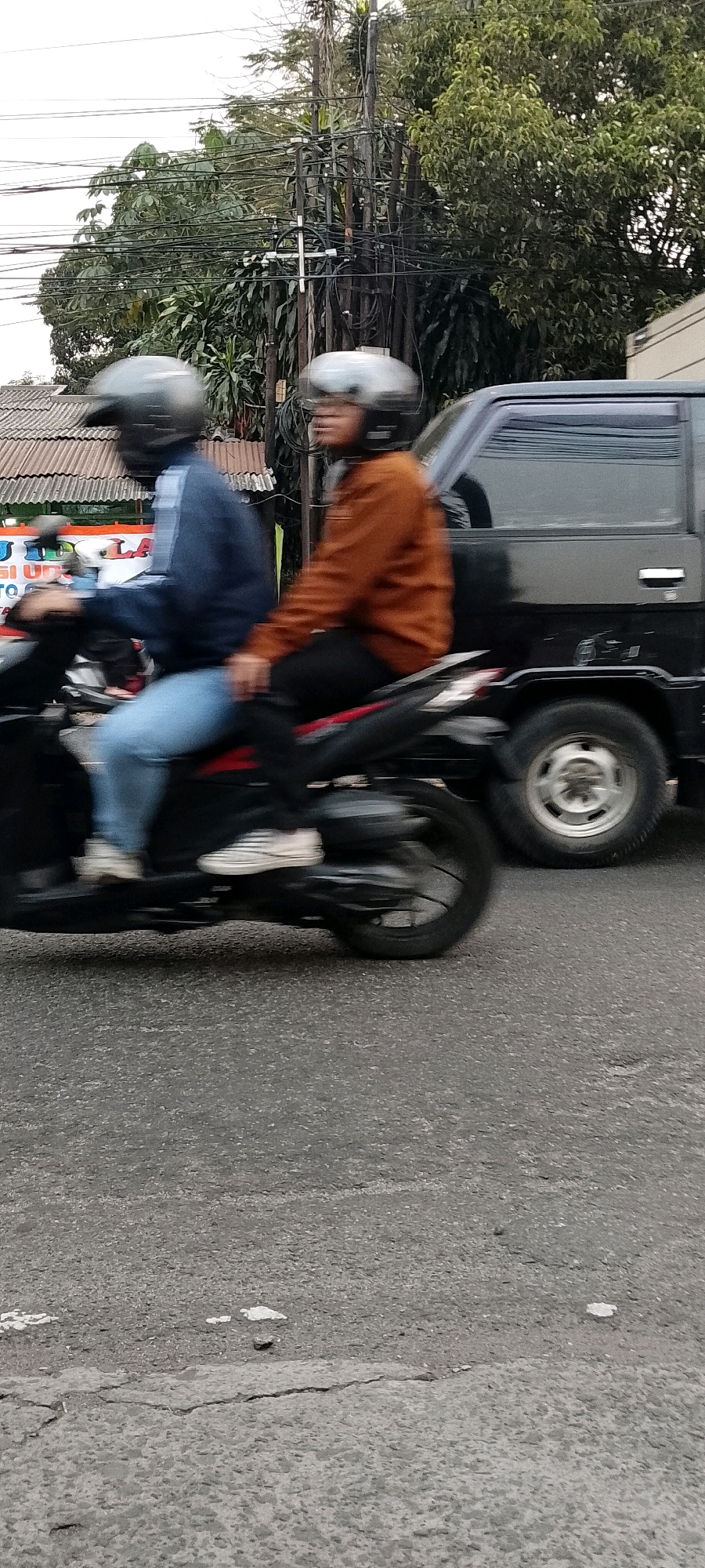 The image depicts two individuals riding a motorcycle on a street, likely in Depok, Indonesia. The person in the front is wearing a dark blue jacket and light blue jeans, with a dark helmet. The person in the back is wearing an orange jacket and black pants, also with a dark helmet. They are moving from left to right, indicated by the motion blur. A black van is visible to the right of the motorcycle. In the background, there are trees, utility poles with numerous wires, and a glimpse of a building with a corrugated roof. The road surface appears to be asphalt with some cracks and white markings. The overall scene suggests a typical urban street environment.