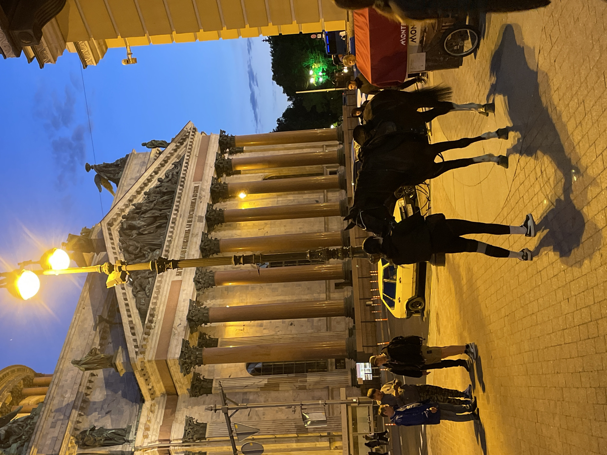 This image depicts an evening scene on a cobblestone street in front of a grand, classical building, likely St. Isaac's Cathedral in Saint Petersburg, Russia. The building features large columns and intricate sculptures under a twilight sky. A streetlamp with two lit globes illuminates the foreground, casting long shadows from a dark horse and several people standing nearby. The horse, saddled and ready, stands centrally in the frame. To the left, a group of four individuals, dressed in casual outdoor attire, are gathered on the sidewalk. A yellow car is parked behind them, and further down the street, orange traffic cones are visible. To the right, a red cart with a visible "MONT" inscription is positioned next to the horse. A person in dark clothing stands near the cart, and another person, partially obscured, is to their right. The overall atmosphere is illuminated by artificial lighting, creating a warm glow on the paved surface.