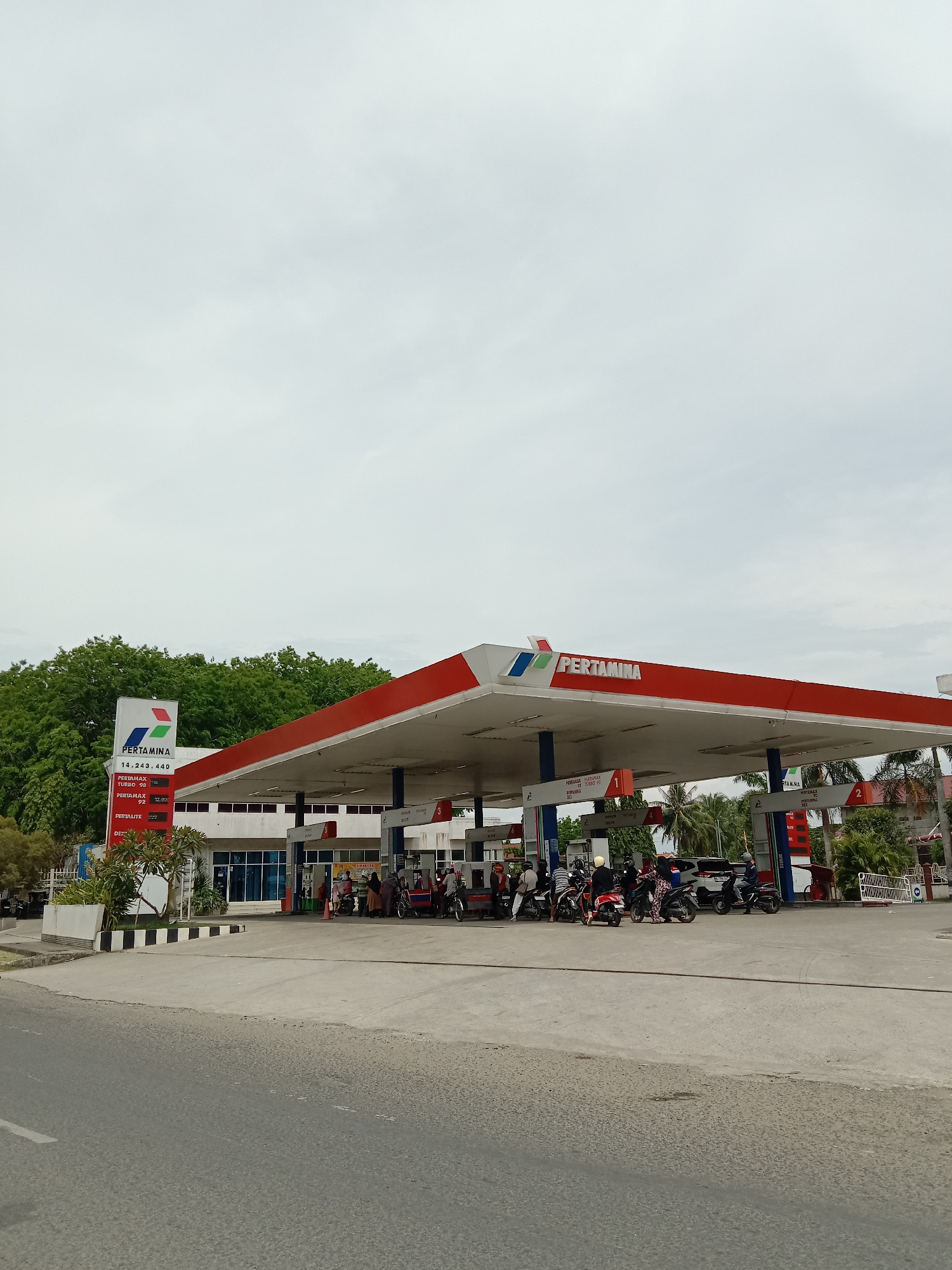 The image depicts a Pertamina gas station located in Lhokseumawe, Indonesia. The station has a large canopy supported by blue pillars with red edges and the Pertamina logo prominently displayed on top. Several motorcycles are lined up at the gas pumps. A price display indicates the cost of different fuel types: Pertamax Turbo 98, Pertamax 92, and Pertalite. The sky is overcast. There is a building to the left of the gas station, and trees are visible in the background. A road is visible in the foreground.
