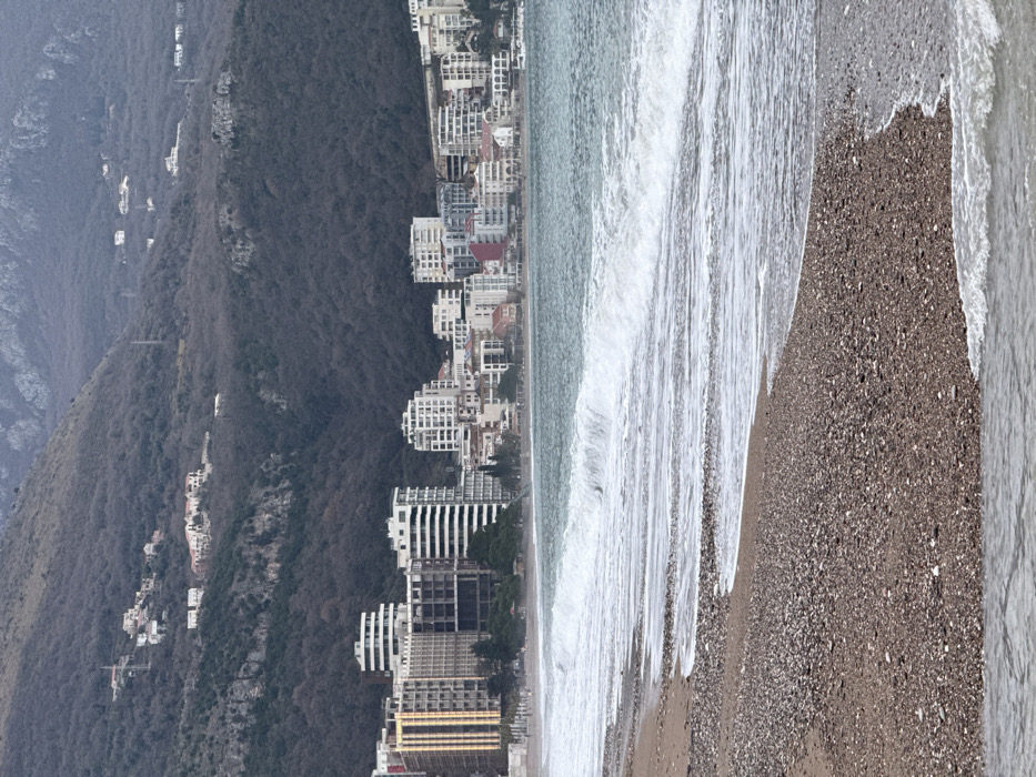 A sideways view of a beachside city nestled against a tree-covered hillside. High-rise buildings line the coast, abutting a narrow strip of beach. White-capped waves crash onto the shore, mixing with the rocky sand. The hillside rises steeply, dotted with buildings, against a muted sky.