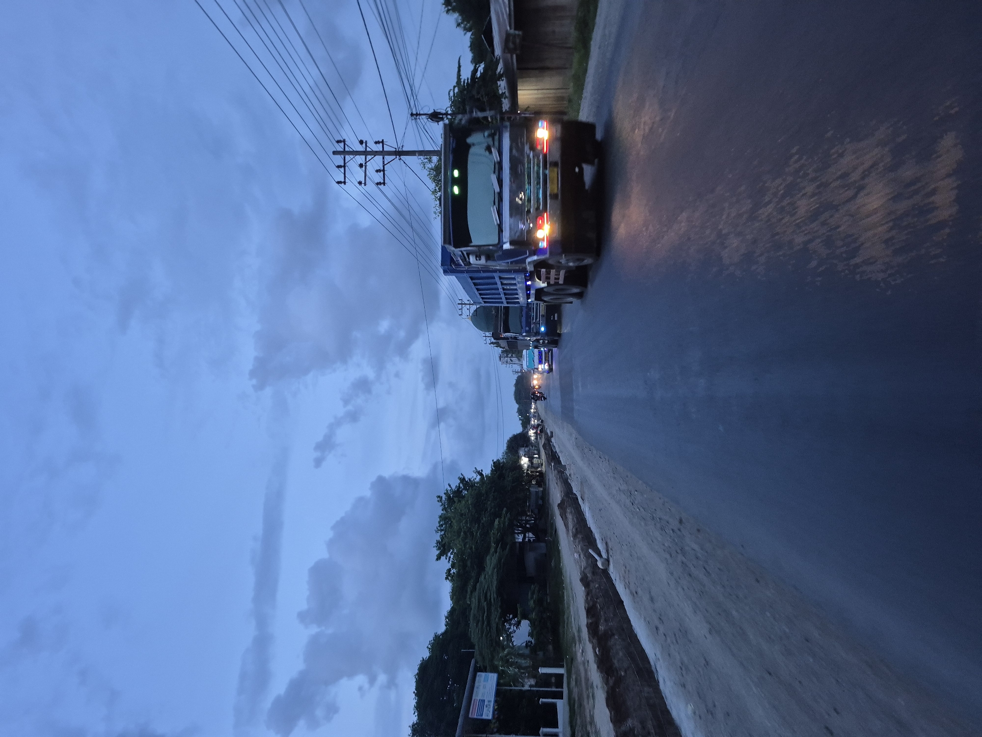 A line of trucks travels along a road in Blangkeude, Indonesia, at dusk. Overhead power lines are visible above the road. Trees line the side of the road, and a ditch runs along the edge. The sky is cloudy.