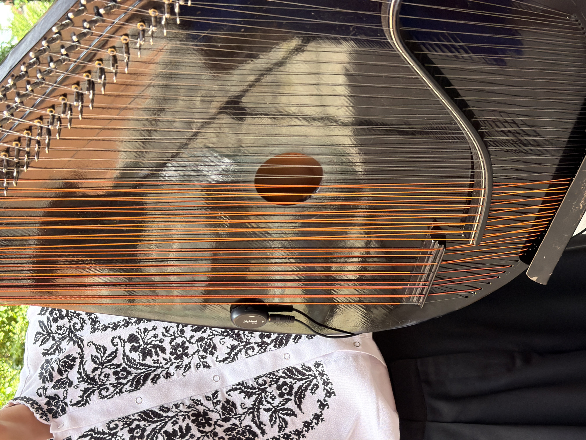 A close-up, vertical, medium shot shows a person wearing a white shirt with black embroidery and black pants, standing next to a large, dark-colored musical instrument with many strings. The instrument is black with a glossy finish and has numerous metal strings running vertically across its surface. Some of the strings are a bright orange color, while others appear to be silver or gold. The shirt is adorned with intricate black floral embroidery, particularly around the collar and chest area. A small black microphone is attached to the instrument, with a cable running from it. The background is softly blurred, with hints of green foliage and a brown textured surface, suggesting an outdoor or natural setting. The image captures a detailed view of the instrument's strings and the textured fabric of the shirt.