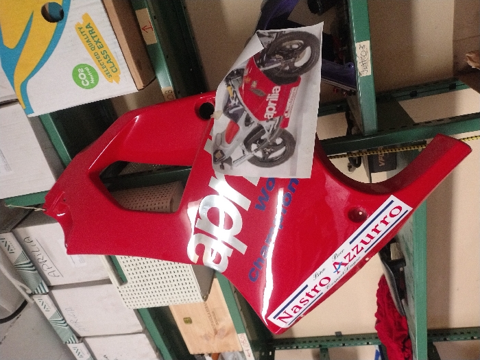 The scene depicts a red Aprilia motorcycle fairing suspended from a green metal storage shelf in what appears to be a storage room or workshop located in Fentange, Luxembourg. The fairing, likely a side panel, features "aprilia" in large silver lettering and "Champion World" in blue. A white sticker with "Nastro Azzurro" and "Birra chiara / Lager premium" in blue and red text is affixed to the lower left portion of the fairing. A color photograph of a red and silver Aprilia motorcycle is taped to the fairing's surface. The fairing is held by a thin green wire.
The surrounding environment consists of green metal shelving units against a light-colored wall. The top shelf holds several white cardboard boxes, some labeled "APRILIA." Below this, a white perforated plastic basket is visible, alongside a large cardboard box marked "CO2 Neutral" and "SELECTED QUALITY CLASS EXTRA." A lower shelf contains a folded red cloth, a black object, and a small wooden box. The shelf below that is labeled "Just_03." No individuals are present in the image.