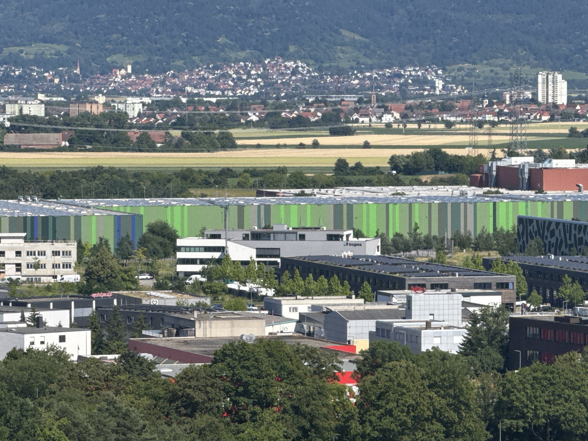 The image shows a panoramic view of a city, likely Mannheim, Germany, with a distinct industrial area in the foreground and a residential area and hills in the background. In the immediate foreground, there is a large, long building with a striking green and white striped facade, possibly a warehouse or distribution center. To the right of this green building, there is a dark, modern-looking building with an abstract pattern on its exterior.
Moving further into the scene, a cluster of various commercial or industrial buildings is visible. These range in color from white and grey to dark grey and black, with some featuring red accents. One building has a prominent red logo that appears to read "weishaupt" in cursive script. Another building has a heart symbol visible on its facade. There are also several vehicles, including vans and cars, parked in the vicinity of these buildings, and some people can be observed near the entrances.
Beyond the industrial zone, the landscape transitions to fields of yellow and green crops, indicative of agricultural land. In the distance, a densely populated residential area with numerous houses and buildings, many with red-tiled roofs, spreads across the hills. Several church steeples and taller buildings can be seen interspersed within the urban landscape. The background is dominated by lush green, tree-covered hills, suggesting a natural backdrop to the city. The overall impression is one of a functional industrial zone coexisting with a vibrant urban center and natural surroundings.