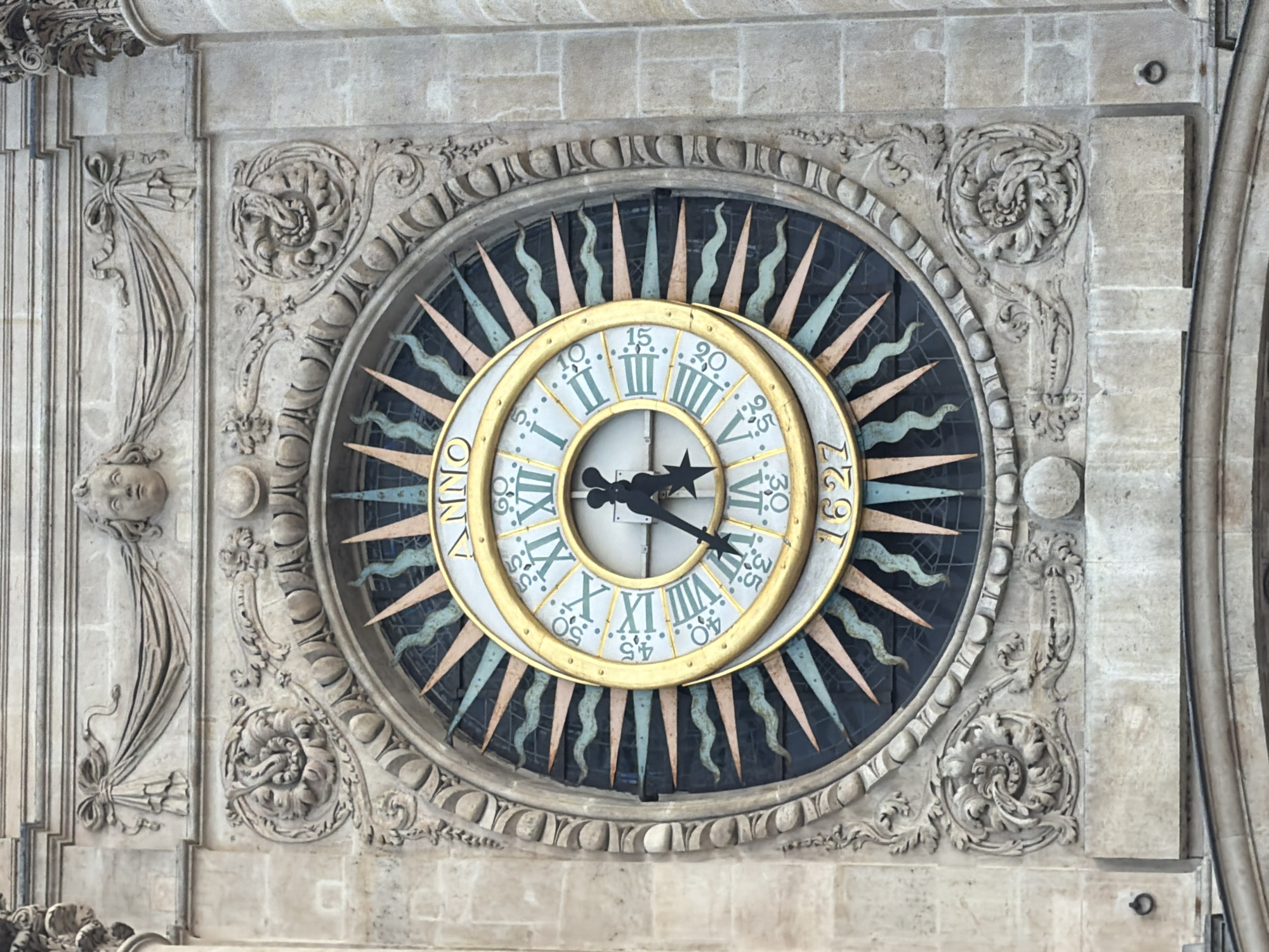 The image shows an old clock on a stone facade. The clock face is decorated with Roman numerals, and the inscription "ANNO 1627" is visible. The clock is surrounded by ornate carvings in the stone. The clock appears to be part of a building. The building is located in Paris, France.