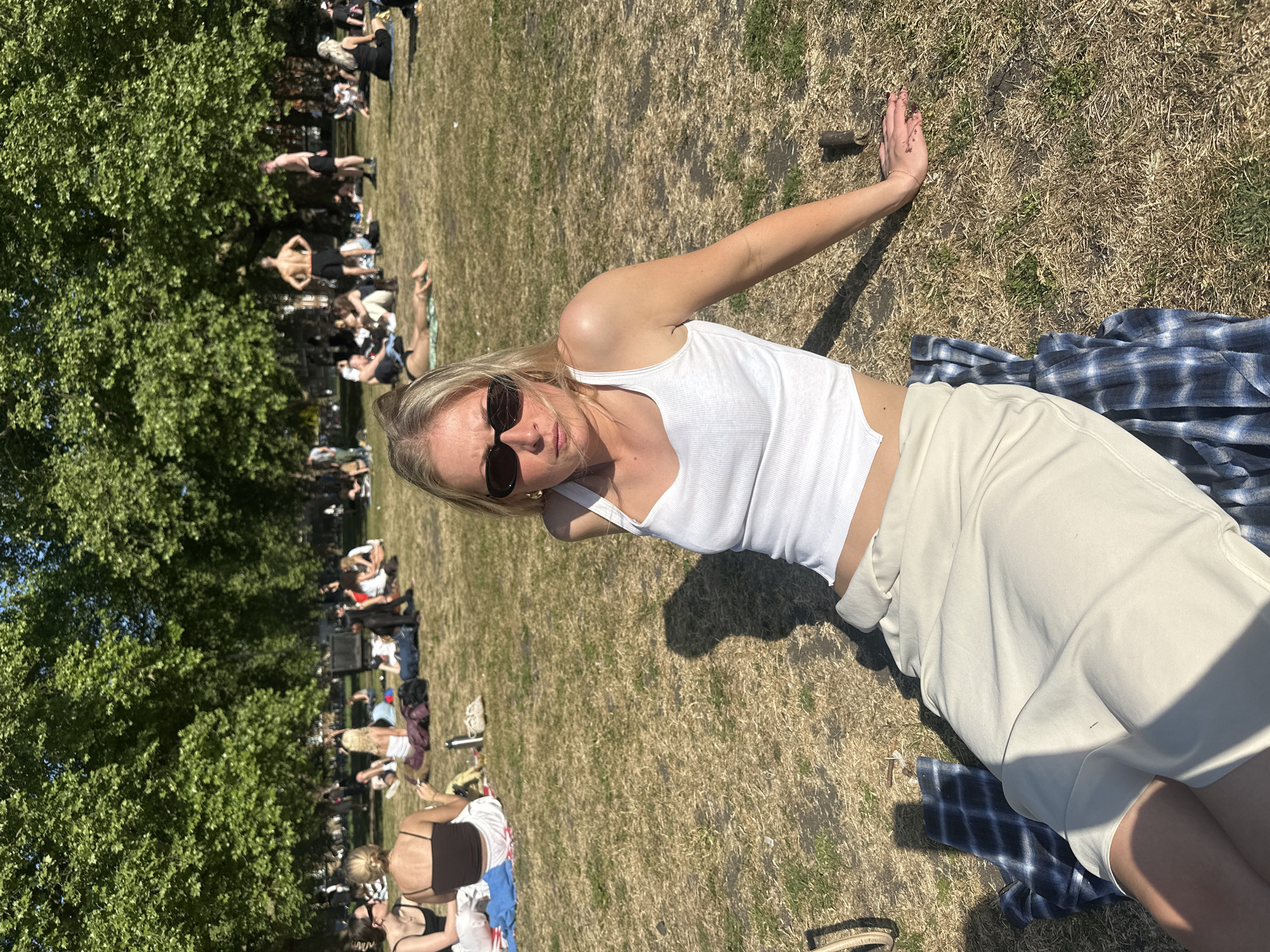 A woman is lying on a blue plaid blanket in a park. She is wearing a white tank top and cream-colored shorts, and sunglasses. She has her right arm outstretched and her hand resting on the grass. In the background, there are people sitting and lying on the grass. There are trees on the left side of the image. The location is likely a park in London, United Kingdom.