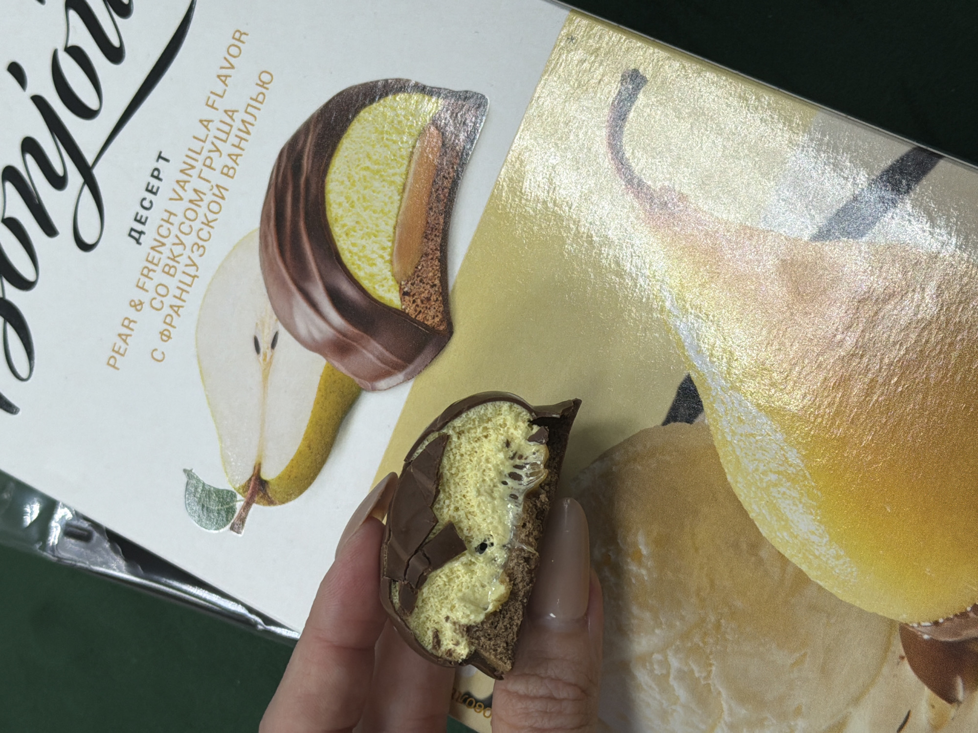 A close-up shot shows a hand holding a half-eaten chocolate-covered dessert with a yellow filling and a caramel-like center. The dessert is placed on top of a box with an image of pears and text that reads "DESSERT PEAR & FRENCH VANILLA FLAVOR WITH FRENCH PEAR VANILLA FLAVOR" in English and Russian. The background is a dark green surface. The scene does not provide any information or context that would allow for identification of a location in the city of Perm, Russia.