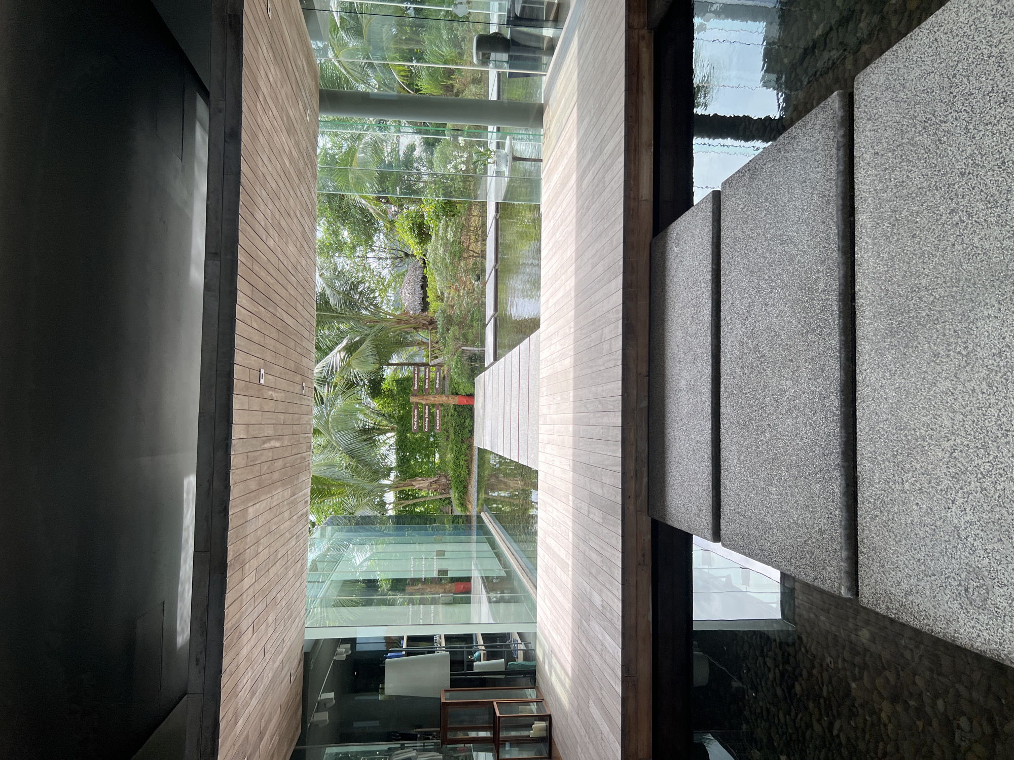 The image depicts an architectural interior leading to an outdoor garden area. The interior features a dark, textured ceiling and a wooden slat ceiling overhead. A large, open space with floor-to-ceiling glass walls reveals a serene outdoor environment.
Dominating the foreground is a wooden walkway that extends across a shallow pool of water. This walkway is composed of horizontal wooden planks. In front of this, a series of three large, grey, rectangular stepping stones are arranged to cross another section of water, which is filled with dark pebbles at the bottom. The stepping stones have a speckled, textured finish.
The outdoor area beyond the glass walls is lush and tropical. Palm trees and other greenery are visible, suggesting a warm climate. A thatched-roof structure, possibly a hut, can be seen in the distance within the garden. Signage is also present in the garden, indicating various stations such as "Chef Garden," "Mushroom Hut," "Warm Cocoon," "Green Reach," and "Birdhouse." The water in the pool reflects the glass walls and the surrounding vegetation.
To the left of the interior space, a glass-enclosed area contains furniture, including a tall white lamp, some shelving units, and decorative items like wooden boxes with candles. The overall impression is one of modern design integrated with a natural, tropical setting, likely a resort or a high-end residence in Pa Khlok, Thailand. There are no people visible in the image.