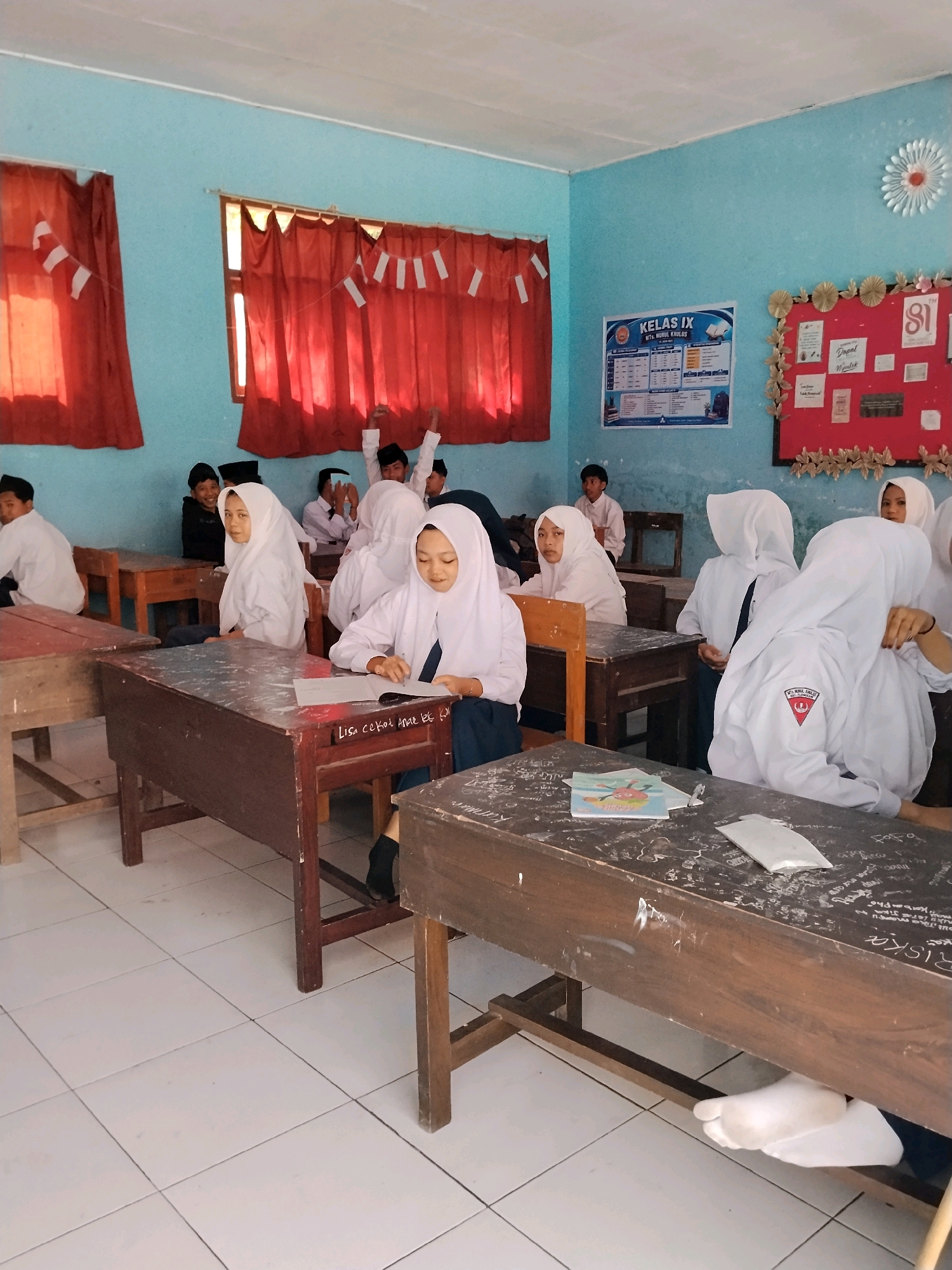 In a classroom in Panggungan, Indonesia, students wearing white uniforms and headscarves are seated at wooden desks. The walls are painted a light blue, with red curtains hanging from the windows. Decorations, including Indonesian flags and a flower made of paper, adorn the walls. A boy in the back raises his hand. A girl in the foreground is reading a book. Another student in the foreground has their feet propped up on a lower shelf of their desk. The floor is tiled with white squares.